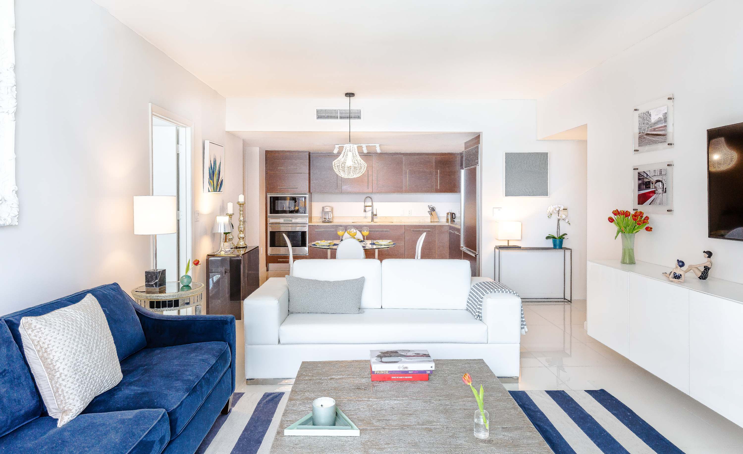 A spacious open-concept living area with white tile floors, a blue sofa, a white sofa, and a rustic wooden coffee table. The modern kitchen with dark wood cabinetry and stainless steel appliances is visible in the background.