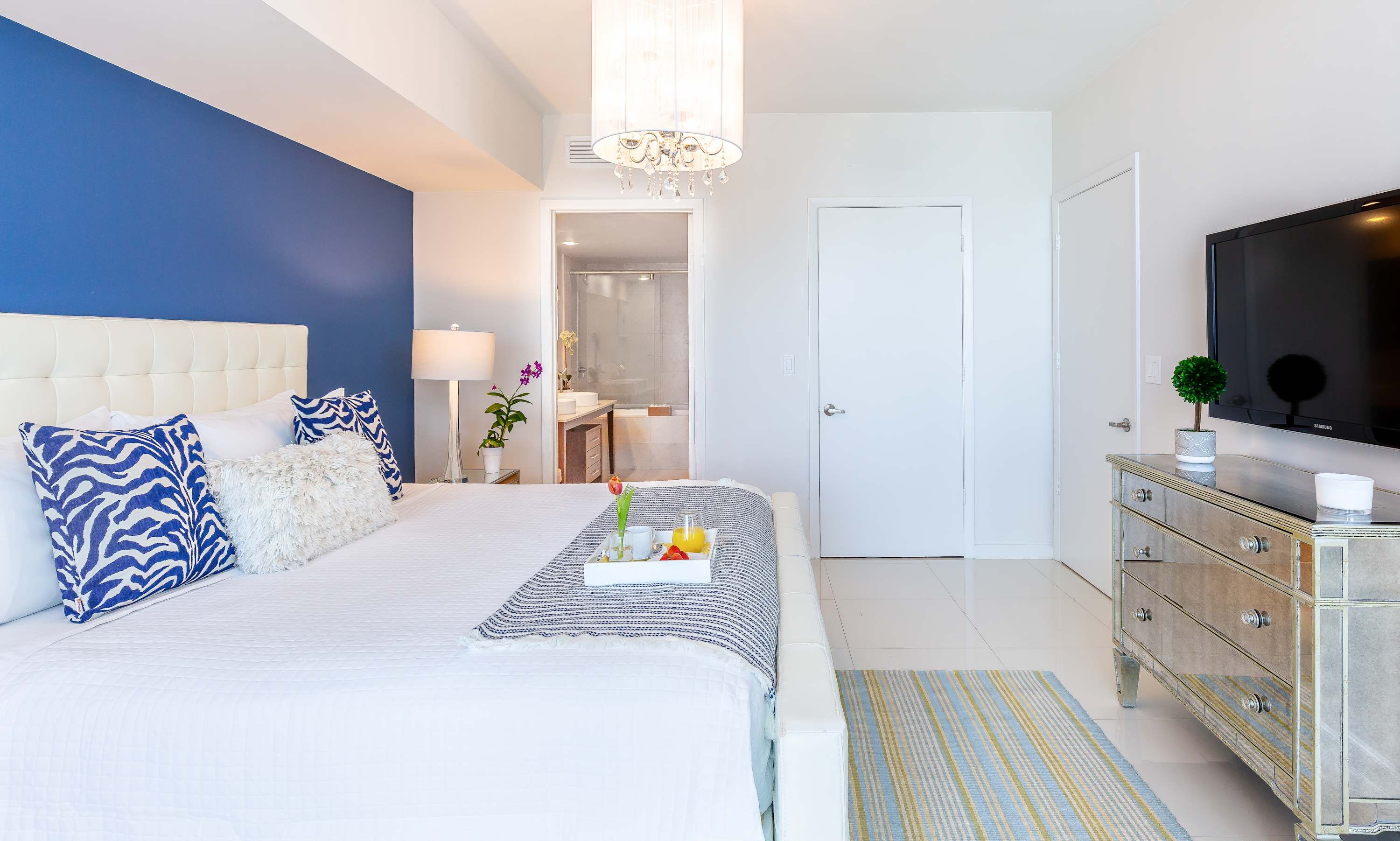 A bright bedroom with white tile floors and a white upholstered bed, featuring a blue accent wall and a mirrored dresser with a flat-screen TV. A doorway leads to the bathroom, revealing a double vanity.