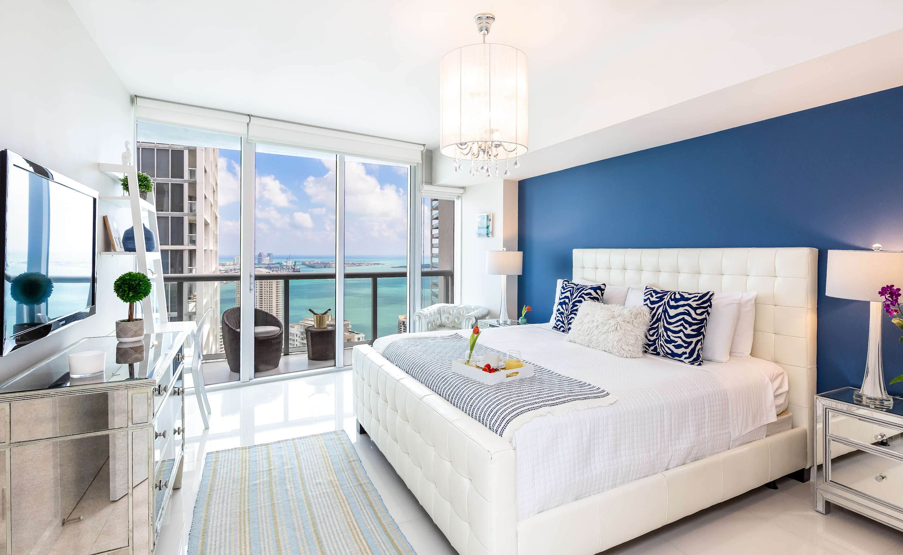 A modern bedroom featuring a white upholstered bed against a bold blue accent wall, white tile floors, and floor-to-ceiling windows with stunning city and water views. A small balcony with seating is visible through the windows.