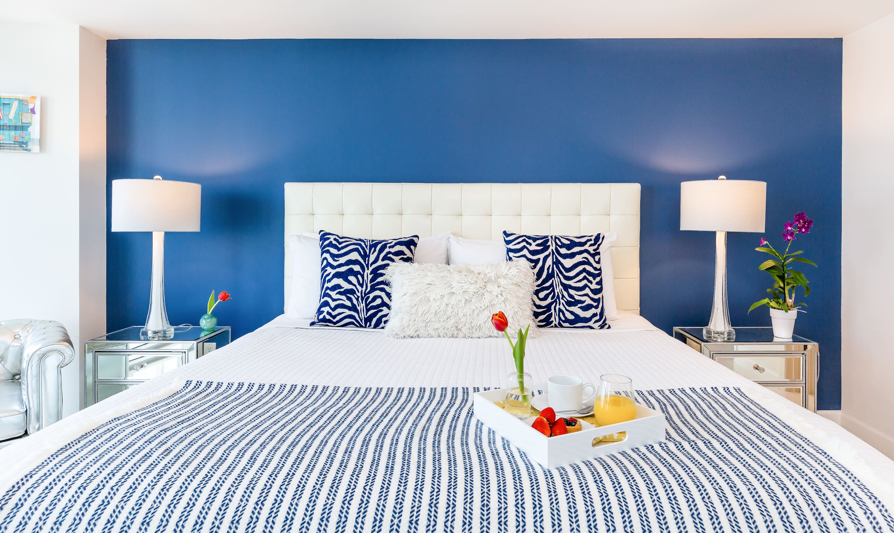 A close-up view of the white upholstered bed in the bedroom, adorned with blue and white striped bedding and decorative pillows, set against a vibrant blue accent wall. Mirrored nightstands flank the bed, each with a lamp.