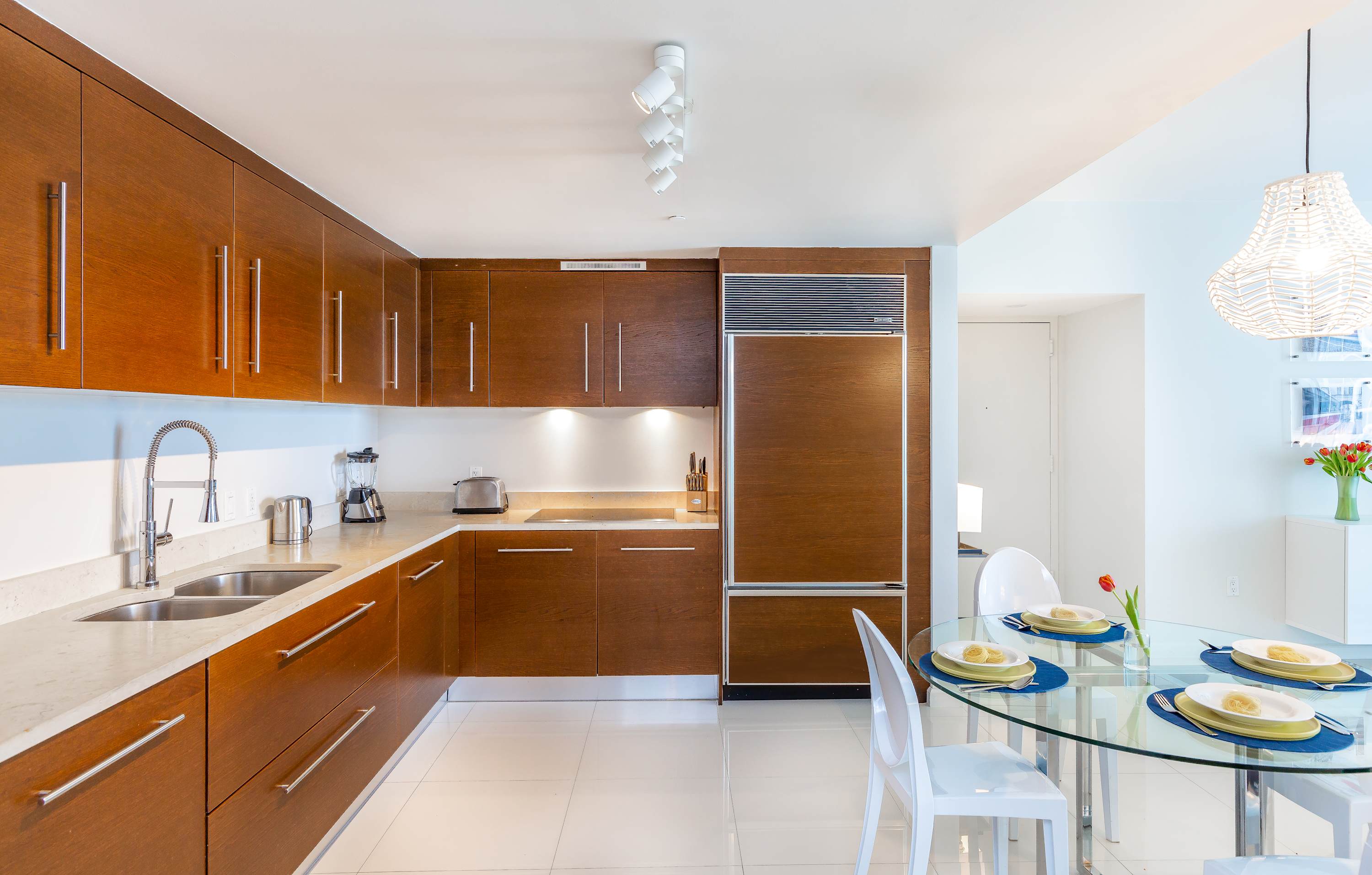 A modern kitchen featuring dark wood cabinetry, light countertops, and stainless steel appliances, including a built-in refrigerator. A glass dining table with white chairs is set for a meal in the foreground.