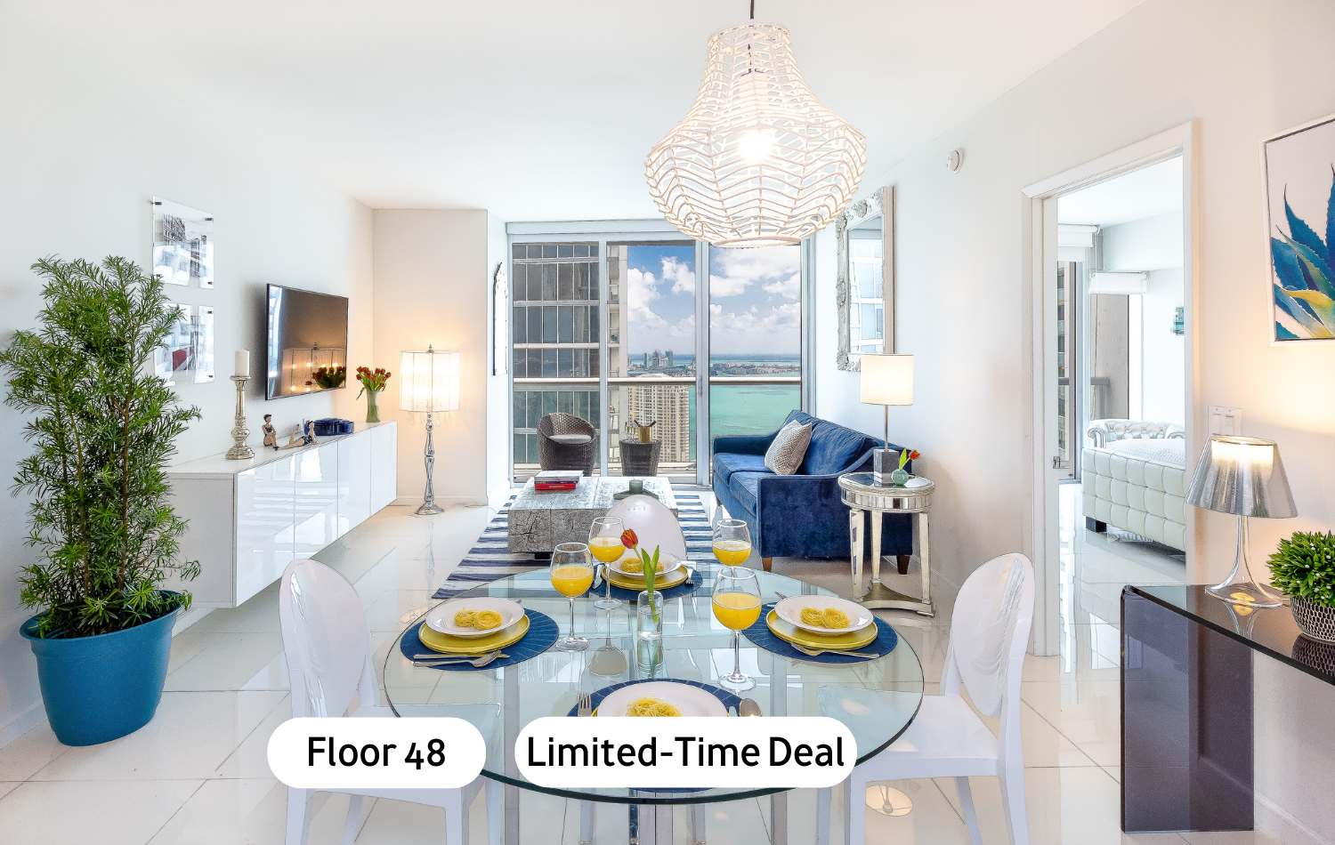 A bright, modern open-concept living and dining area with white tile floors, a blue velvet sofa, a glass dining table with white chairs, and large windows offering city and water views. A large white woven chandelier hangs above the dining table.