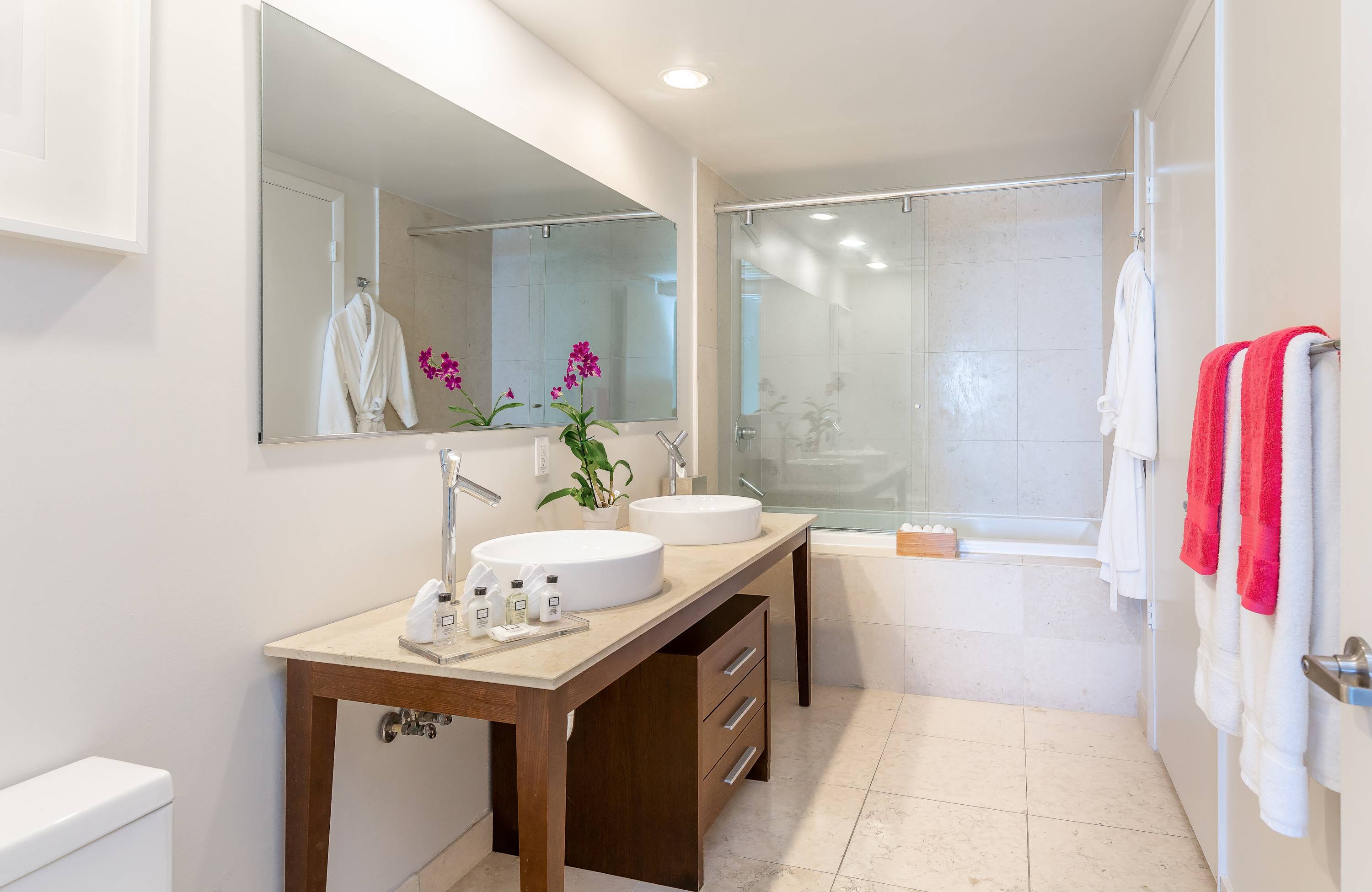 A modern bathroom with light tile floors and walls, featuring a double vanity with vessel sinks and a large mirror. A glass-enclosed shower and bathtub combination is visible in the background.
