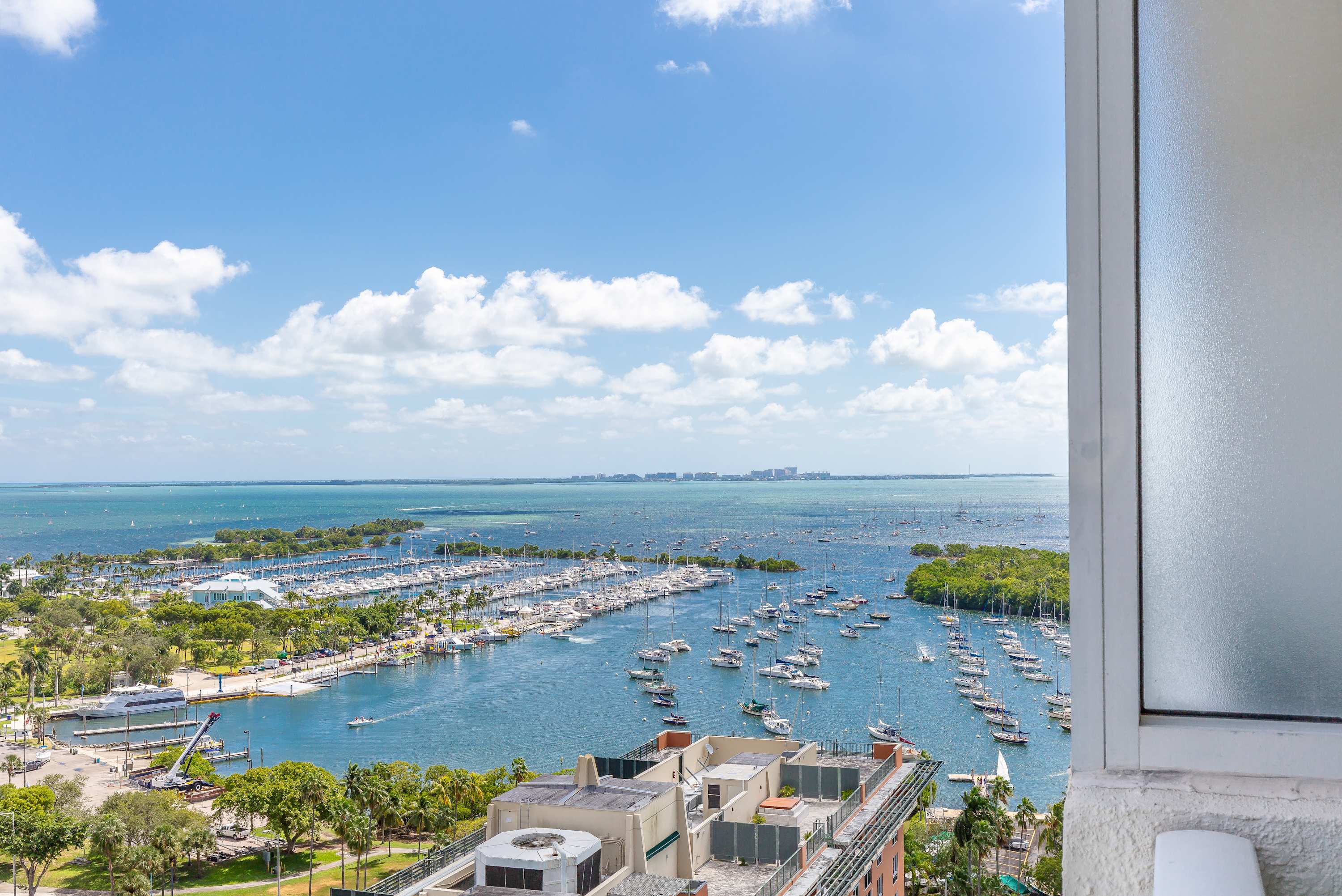 High-angle view from a balcony overlooking a marina filled with numerous boats on a bay, with a clear blue sky and distant cityscapes under fluffy white clouds.