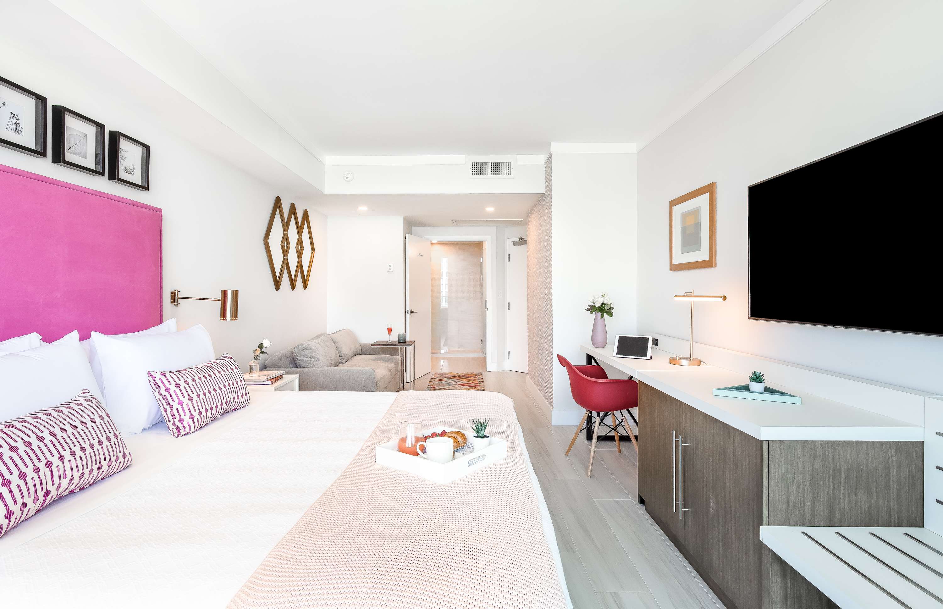 Wide shot of a modern bedroom featuring a large bed with a pink headboard, a grey sofa, and a white desk area with a red chair, leading to a hallway and bathroom entrance.