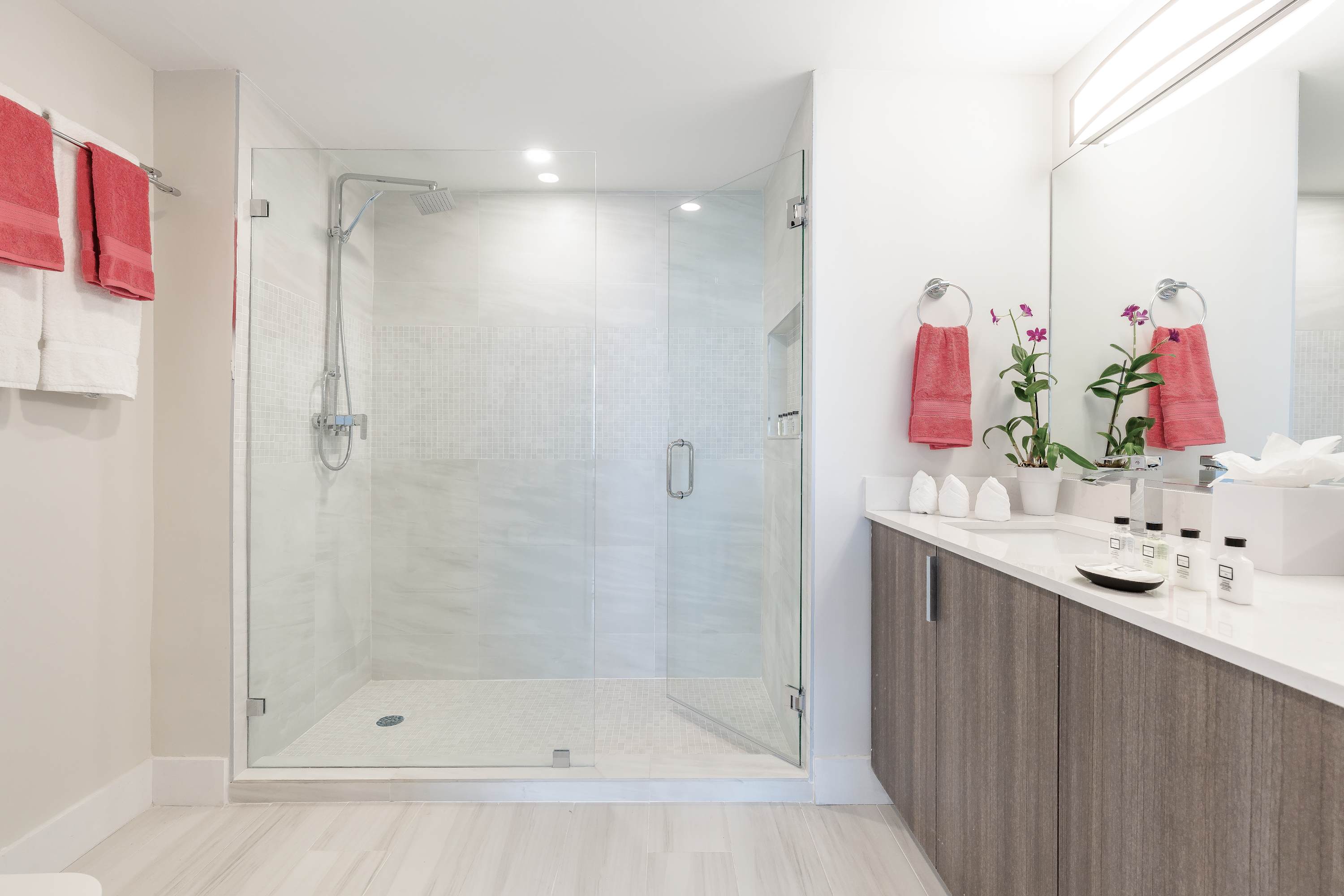 Modern bathroom with a spacious walk-in shower featuring glass doors and light-colored tiles, alongside a vanity with a large mirror and contemporary fixtures.