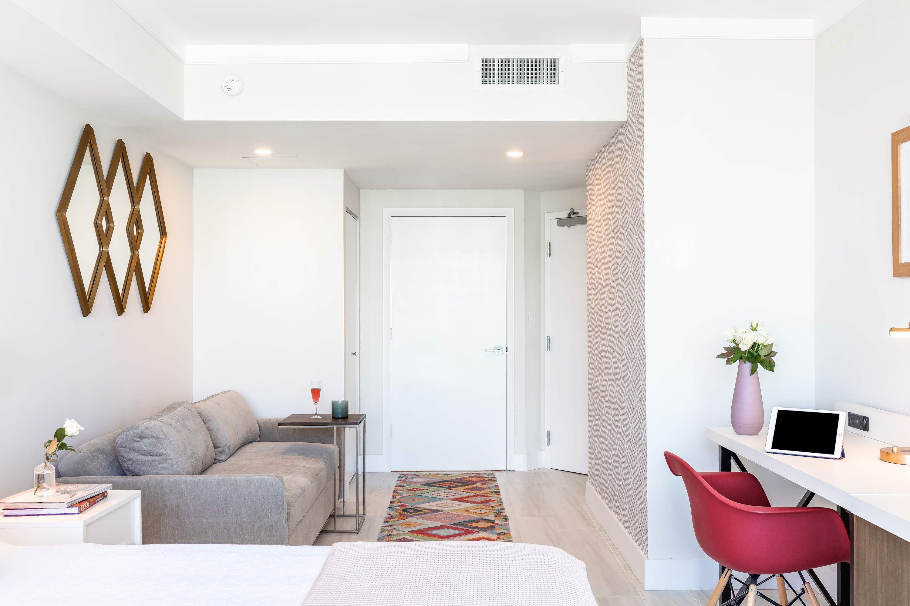 View from the bed towards the entrance, showing a grey sofa, a colorful rug, and decorative wall art, with a glimpse of the hallway and front door.