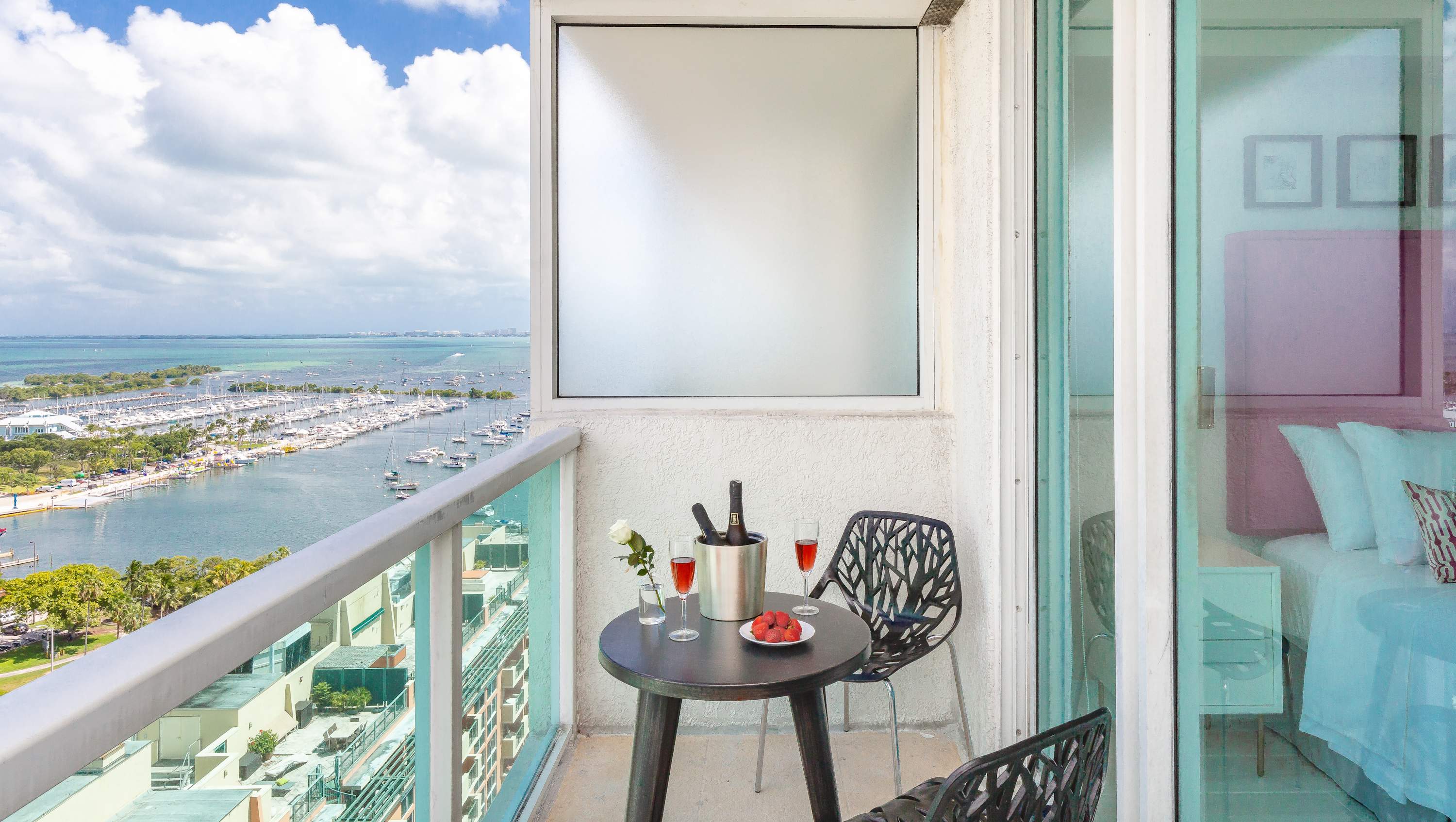 A small private balcony with a round black table and two chairs, set with wine and fruit, offering a stunning panoramic view of a bay filled with boats and distant land.