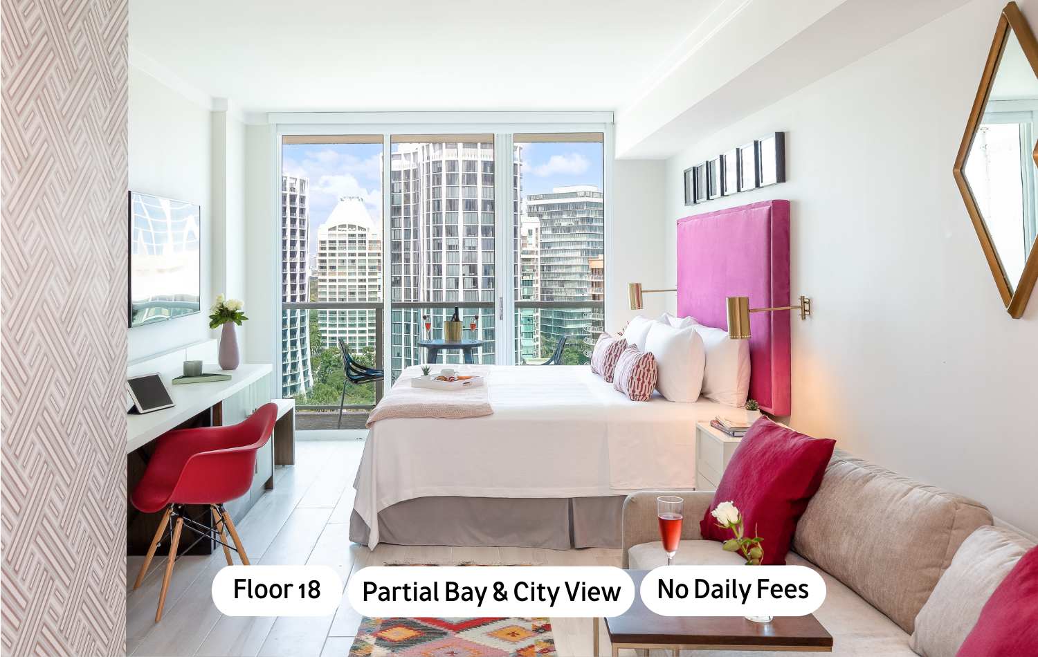 Bright bedroom with a large bed featuring a vibrant pink headboard, a small sofa, a desk area, and a balcony offering partial bay and city views through floor-to-ceiling windows.