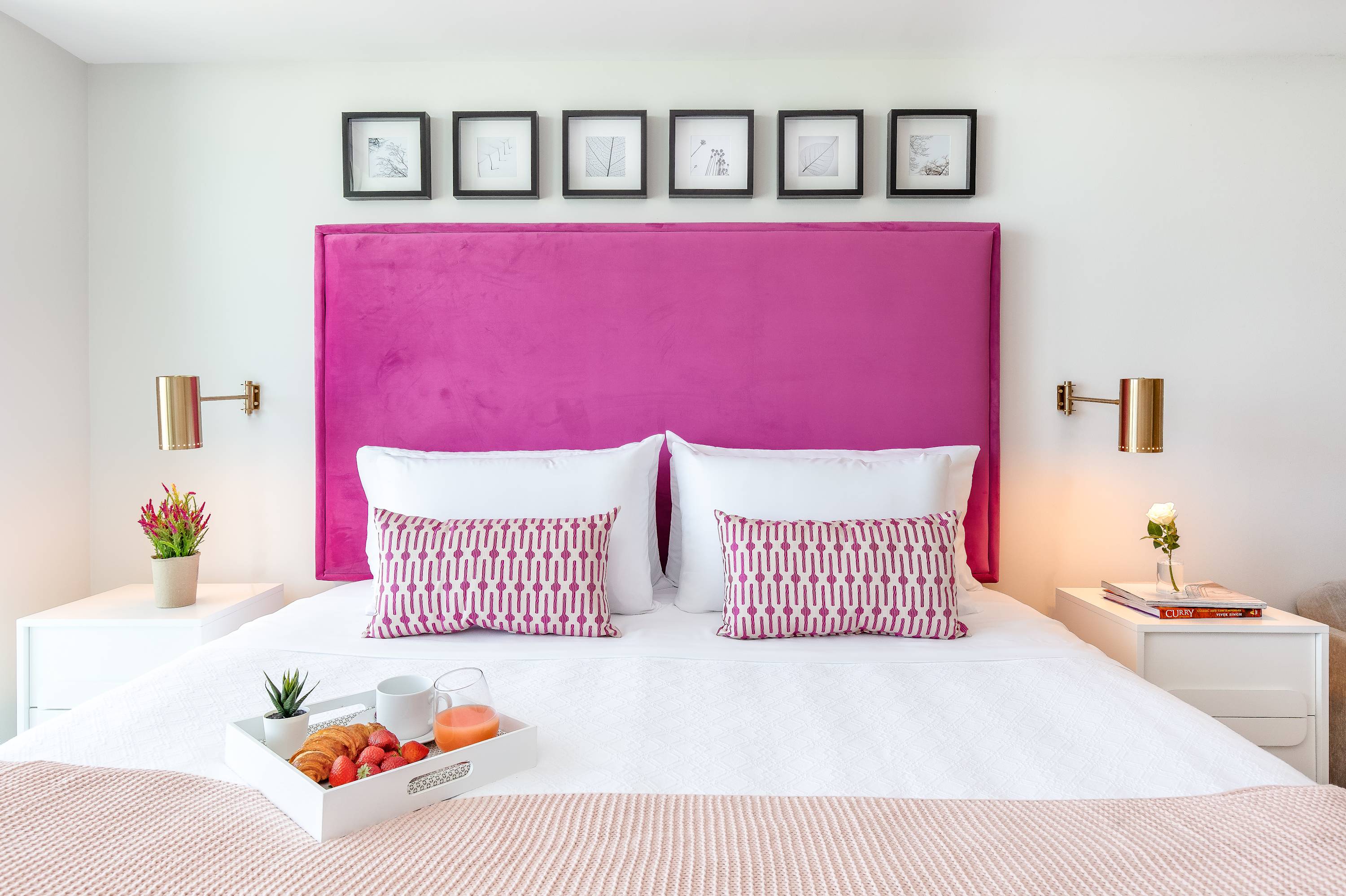 Close-up of a neatly made bed with white linens, a large pink velvet headboard, and two patterned throw pillows, flanked by modern nightstands with gold sconces. A breakfast tray sits on the bed.