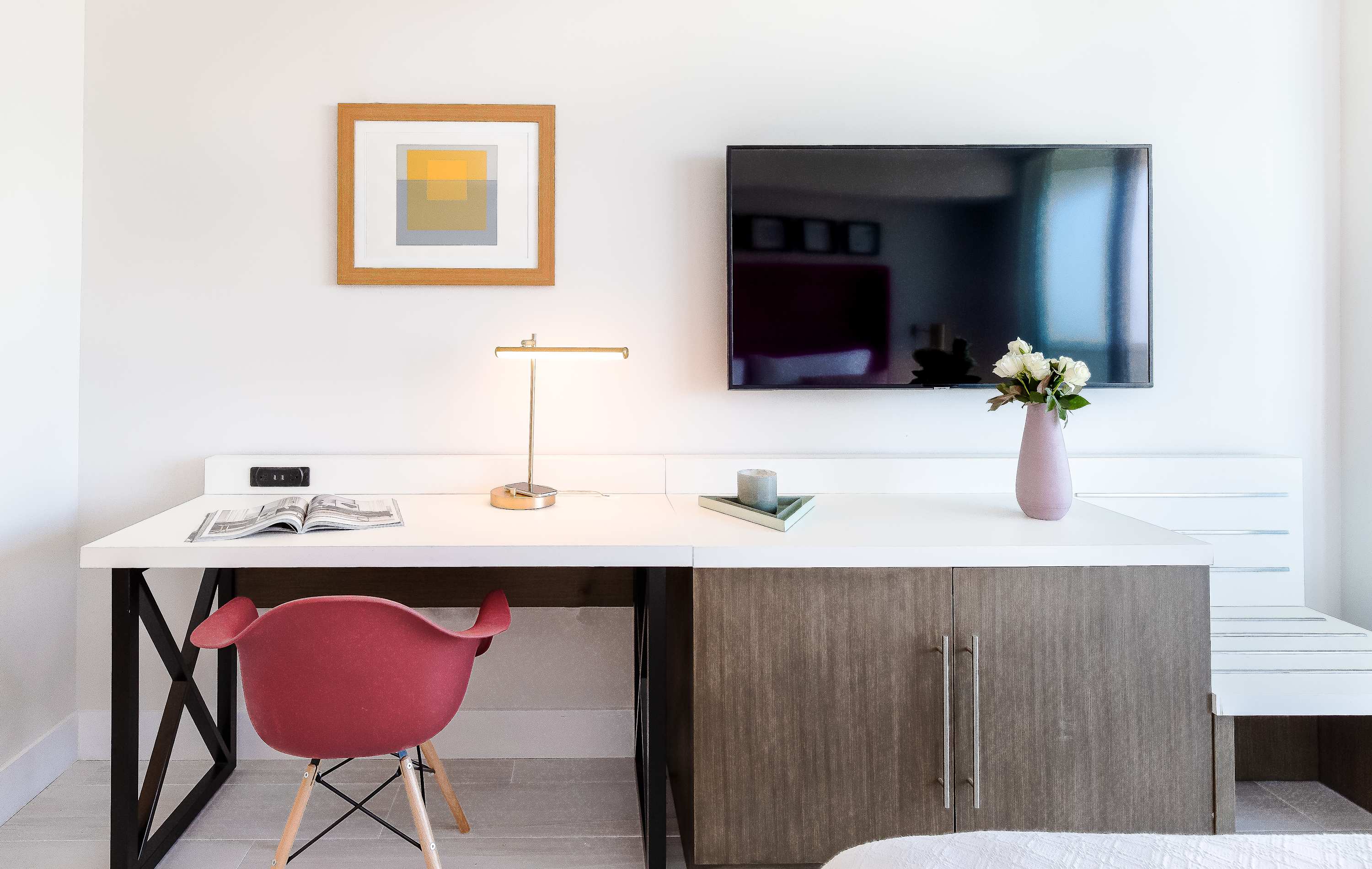 Detailed view of the bedroom's desk area, featuring a white desk with a gold lamp, a red chair, and a wall-mounted flat-screen TV, against a clean white wall with a framed artwork.