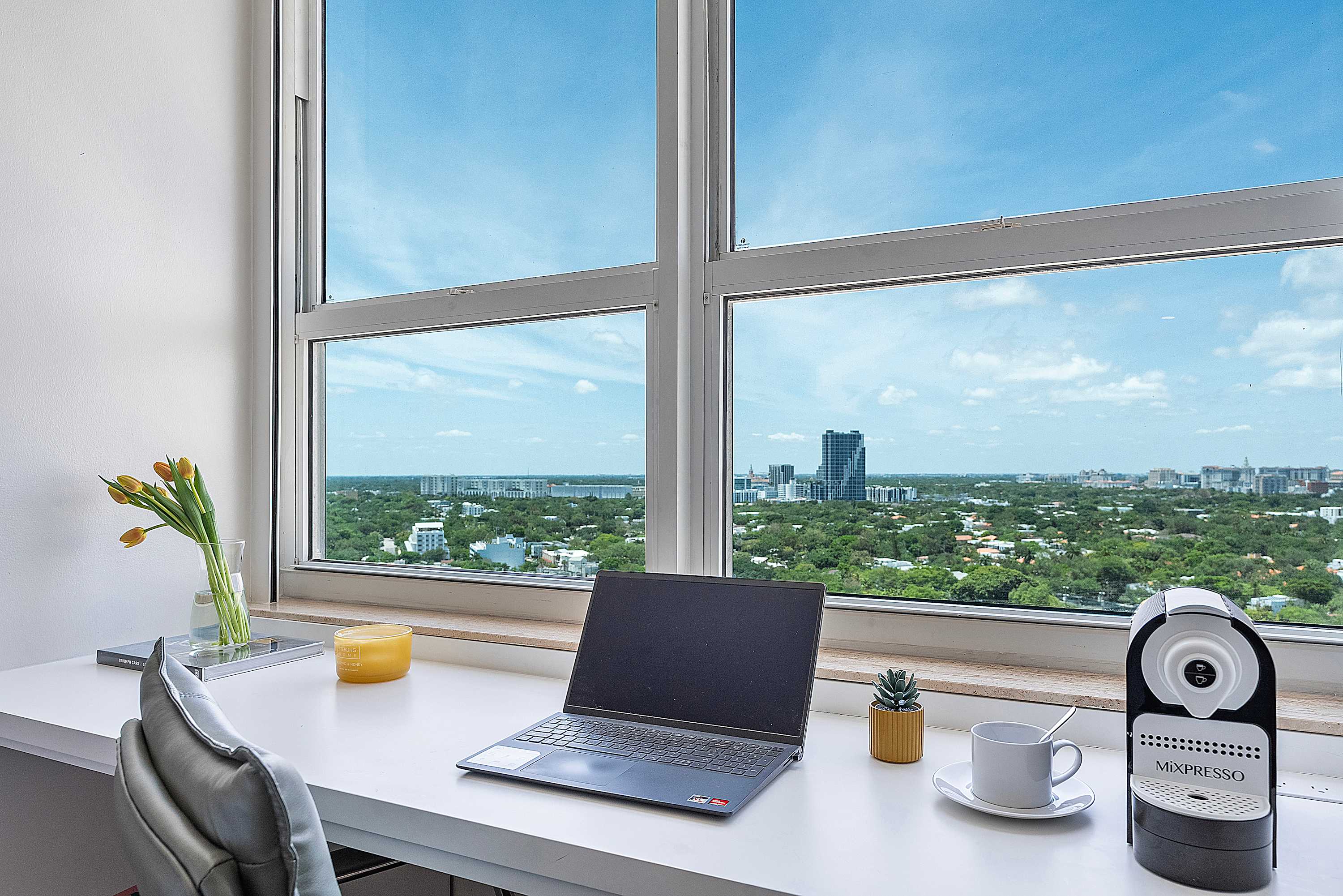 A well-lit workspace by a large window, featuring a laptop, coffee machine, and decorative items, all overlooking a sprawling city and green landscape.
