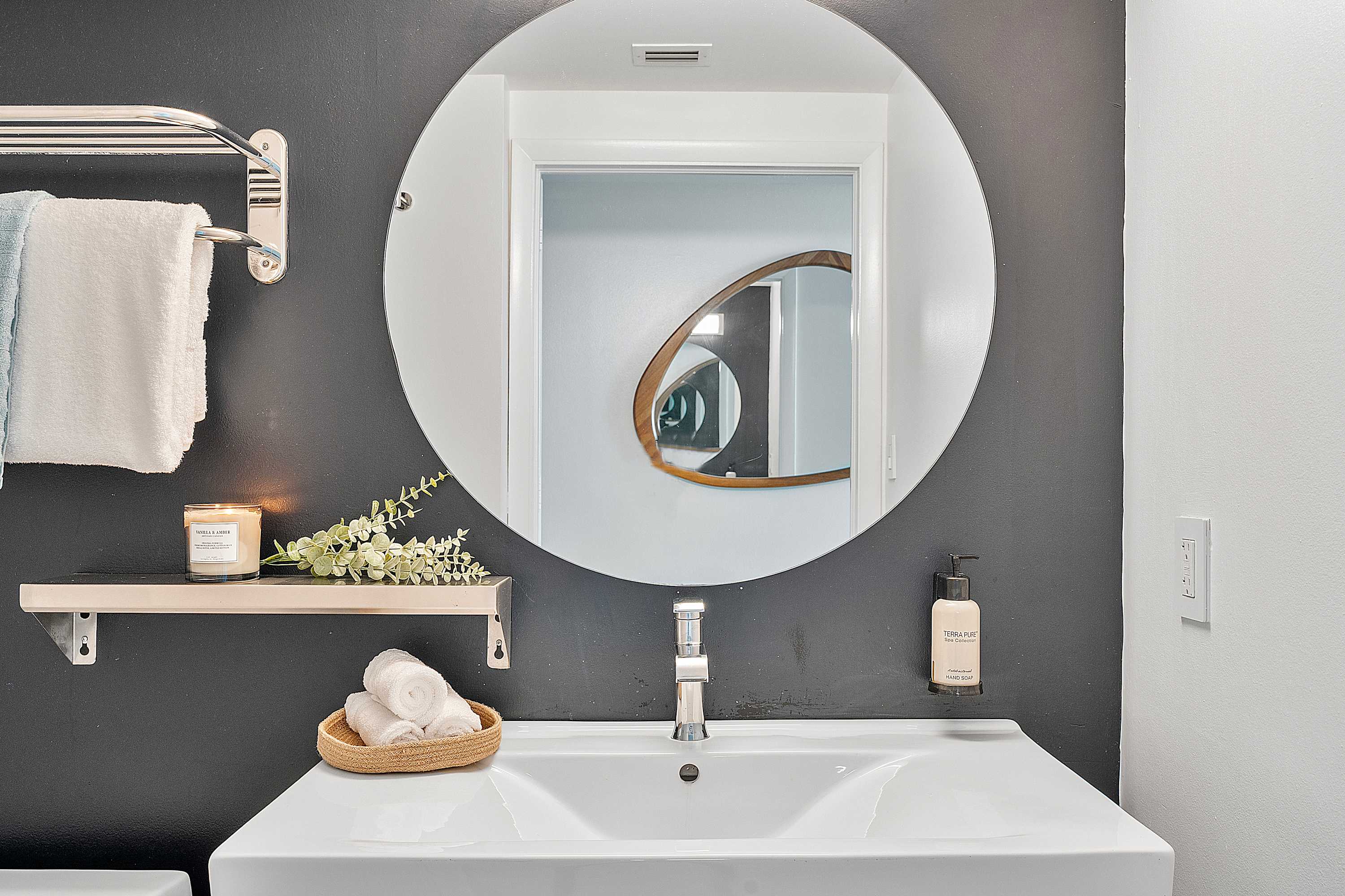 Modern bathroom vanity with a white rectangular sink, a large round mirror, and a dark gray accent wall. A small shelf holds a candle and plant, with fresh towels nearby.