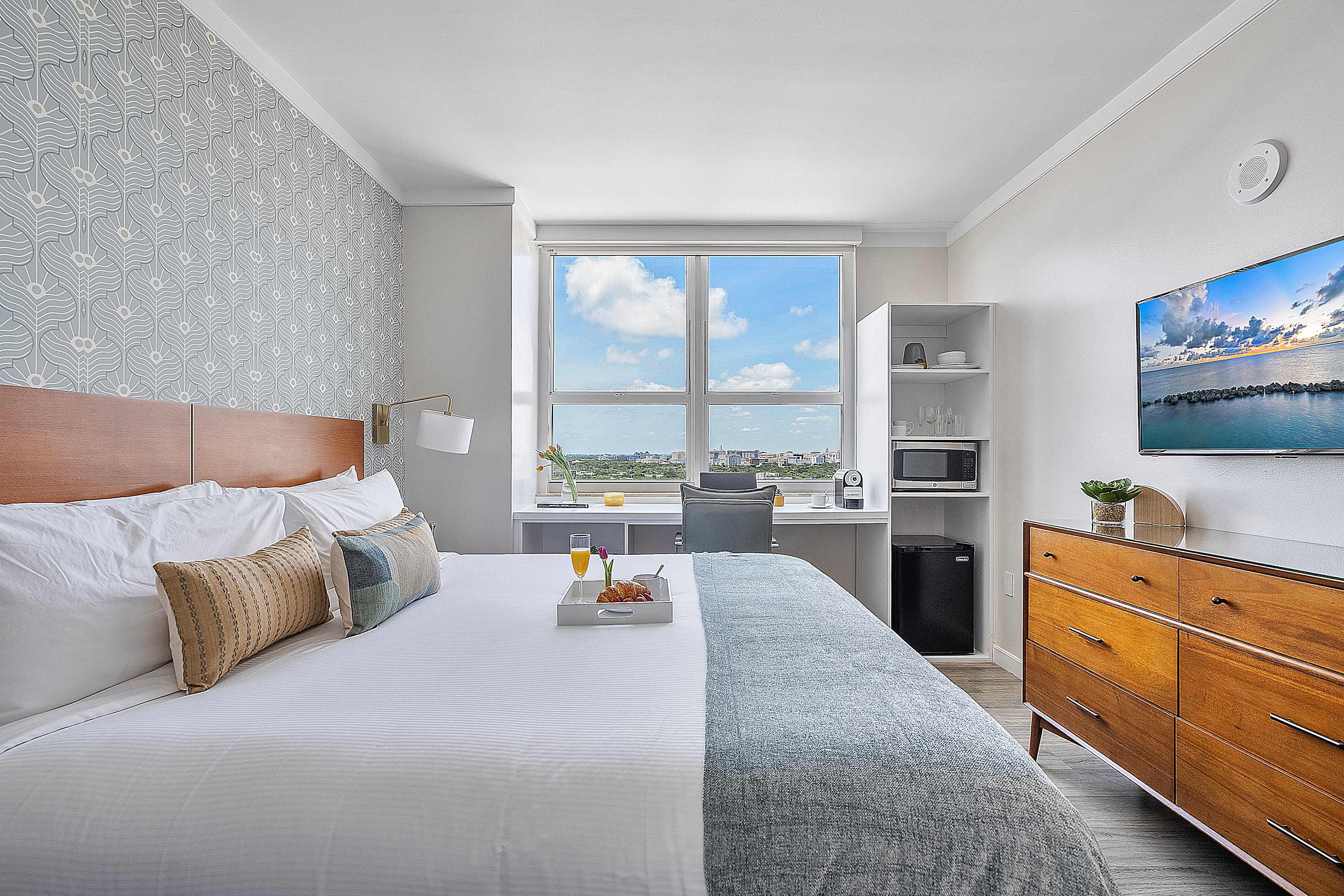 Spacious bedroom with a large bed, a wooden headboard, and a patterned accent wall. It includes a desk by the window, a flat-screen TV, a mini-fridge, and a wooden dresser.