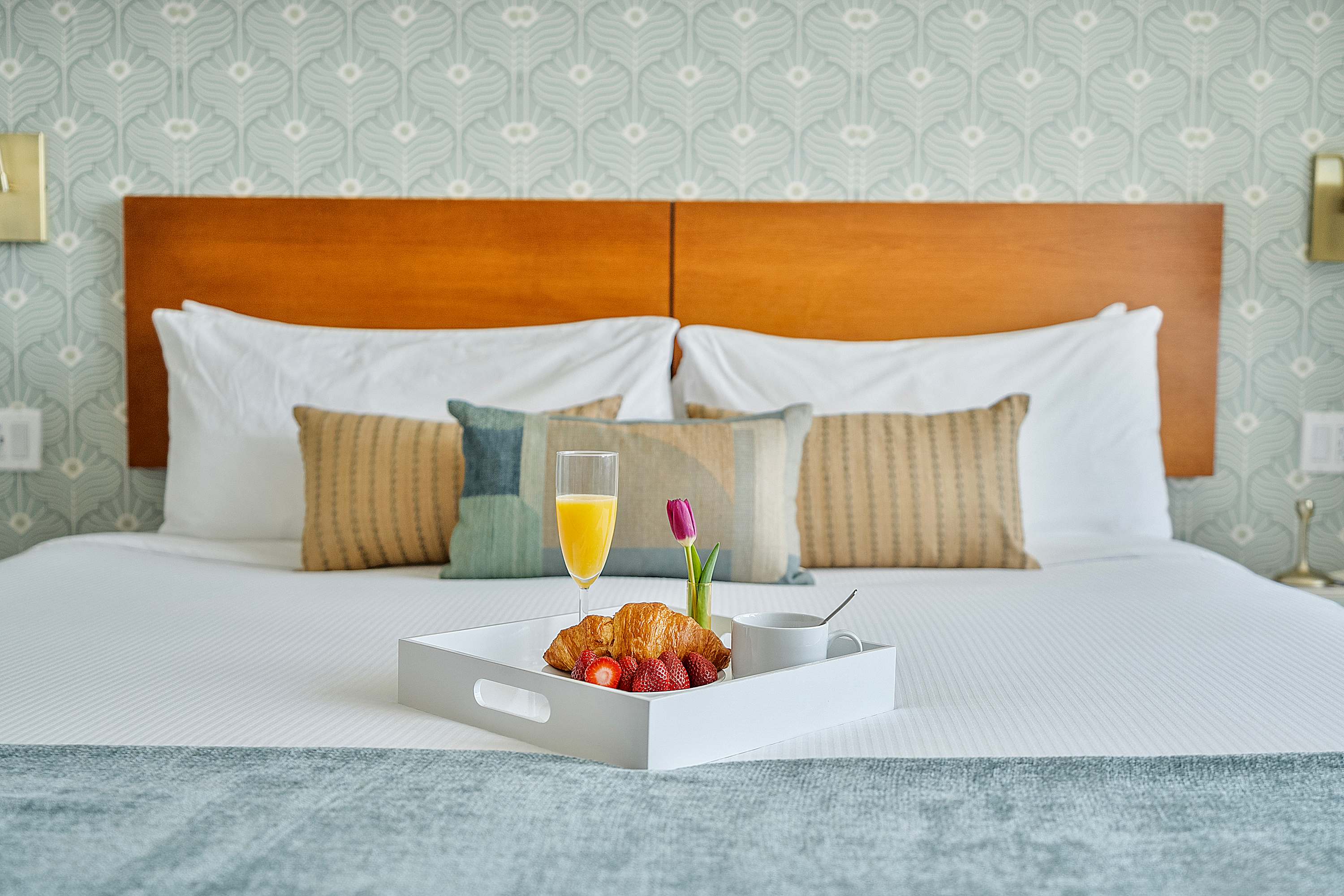 Close-up of a comfortable bed with a wooden headboard and patterned wallpaper, featuring a breakfast tray with orange juice, a croissant, and fresh strawberries.