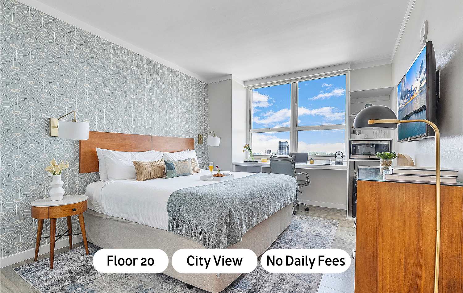 Bright bedroom featuring a large bed with a wooden headboard, patterned accent wall, and a desk by a large window offering a panoramic city view. A flat-screen TV is mounted on the wall.