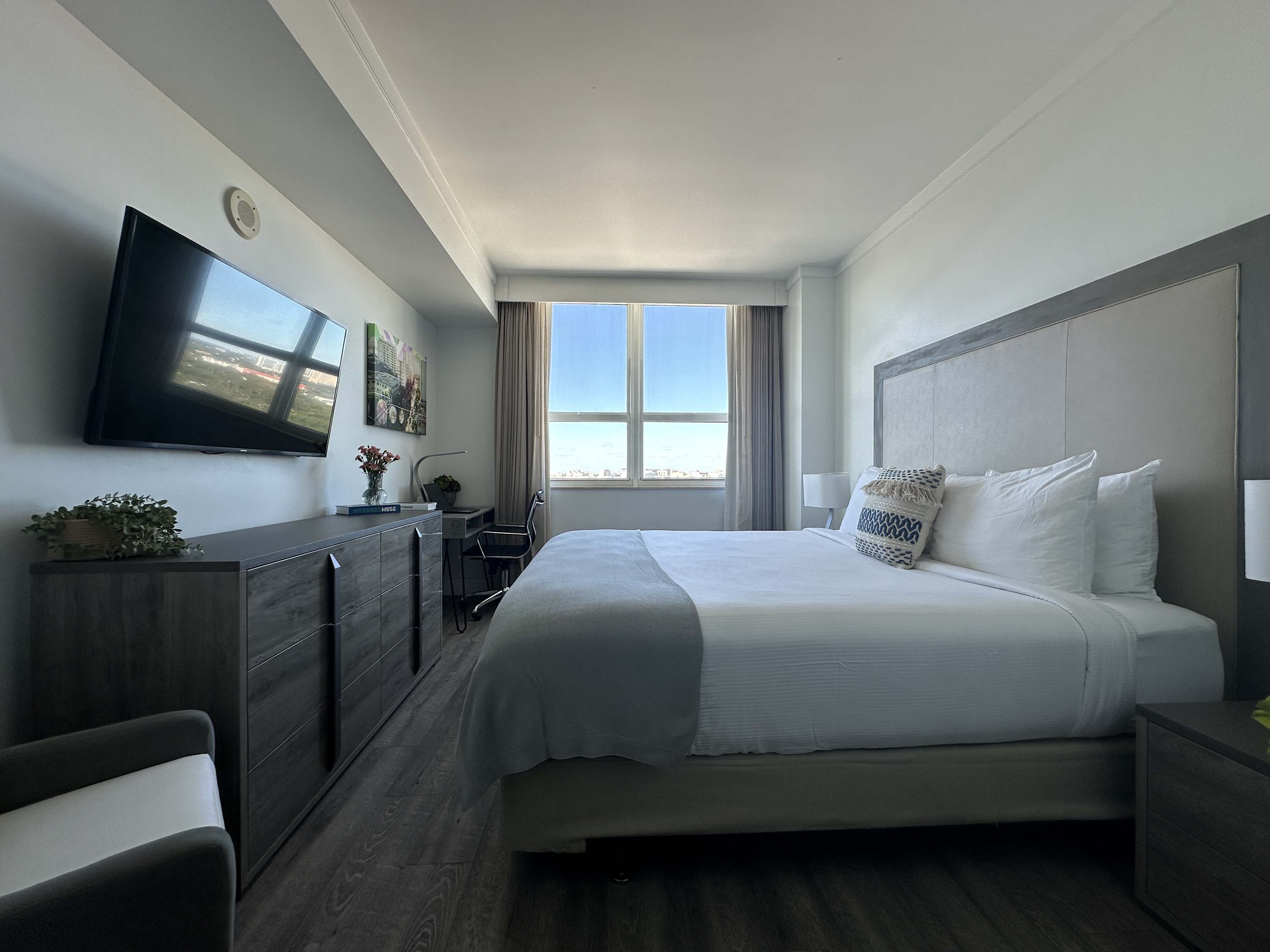 Spacious bedroom with a large white bed, a wall-mounted flat-screen TV, a long wooden dresser, and a dedicated workspace by the window, all with hardwood floors and a city view.