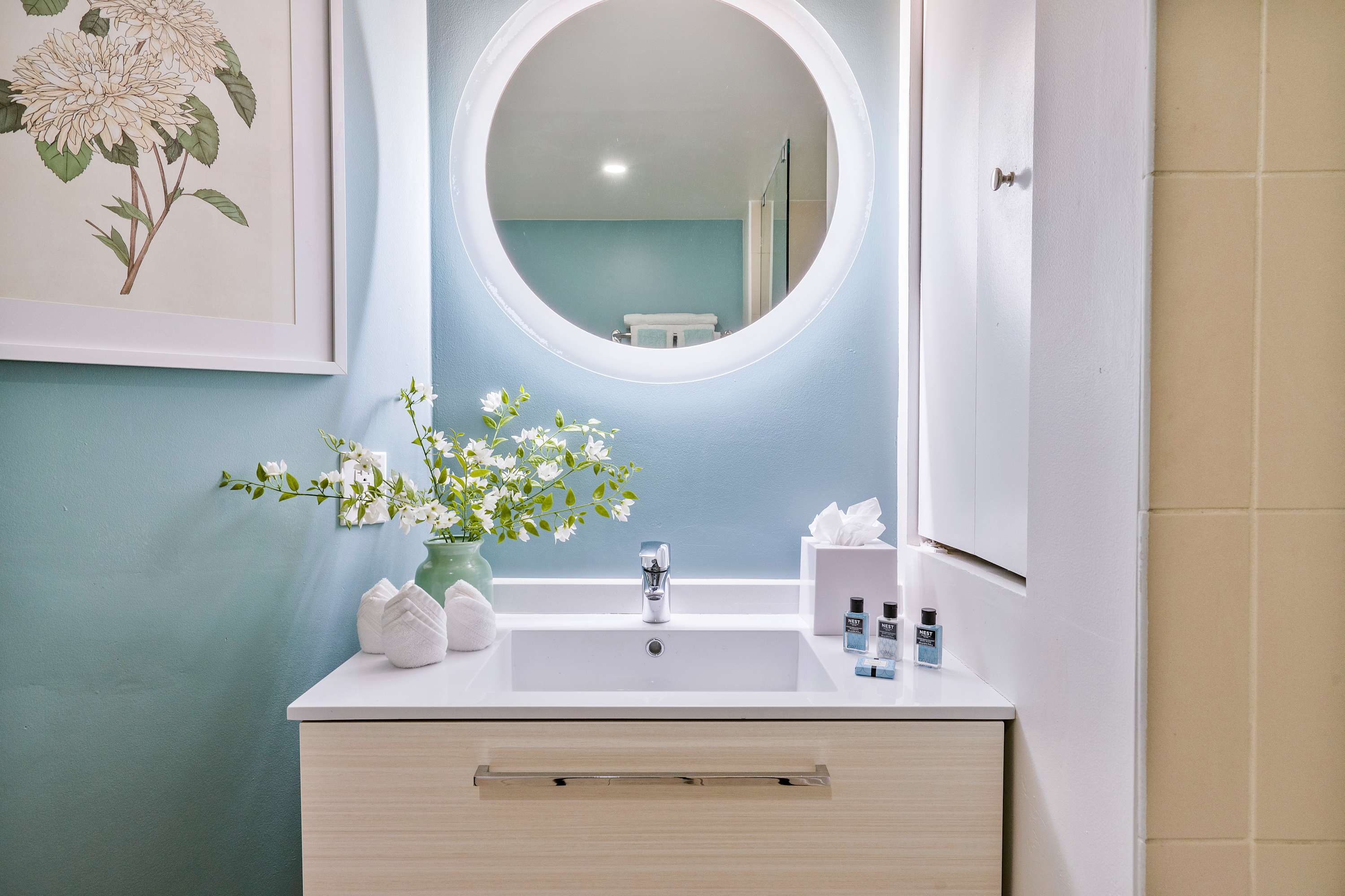 A close-up of the bathroom vanity showcases a white sink, a modern chrome faucet, a large round illuminated mirror, and a vase with white flowers, alongside neatly folded towels and toiletries.