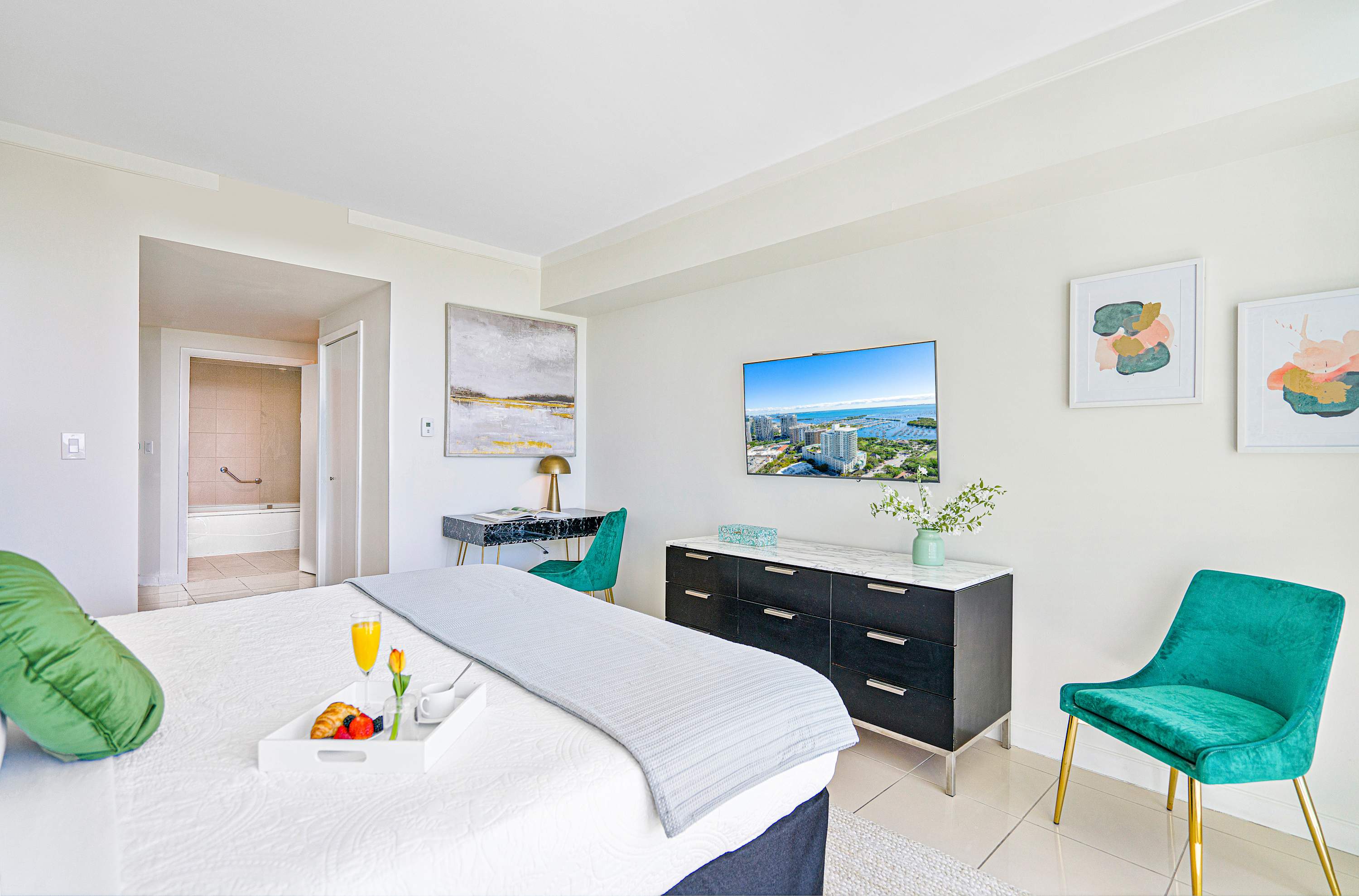 This spacious bedroom includes a large bed, a wall-mounted TV, a black dresser, and a desk area with a teal chair, with an open doorway revealing a glimpse of the bathroom.