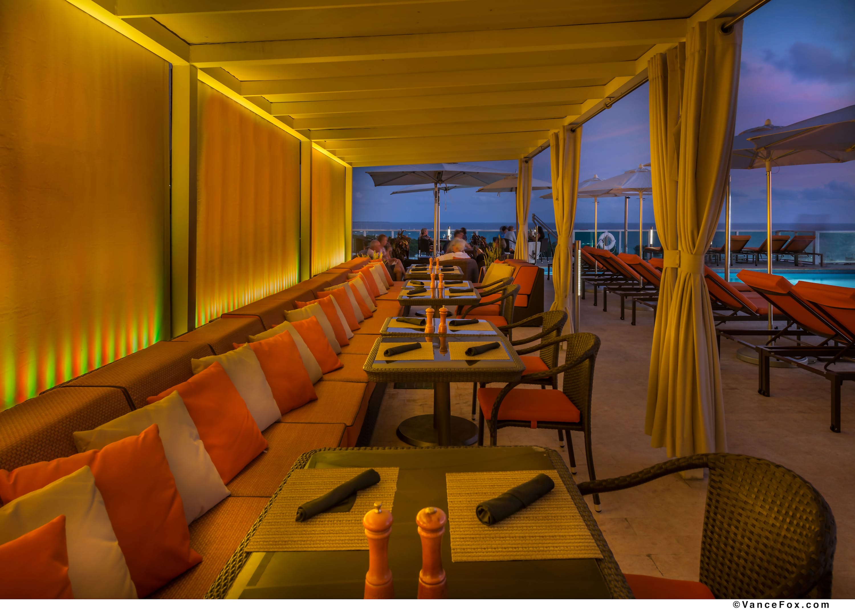 A long, covered outdoor dining area features a row of tables with wicker chairs and orange cushions, set against a vibrant orange and green illuminated wall. Beyond the dining area, a swimming pool is visible with lounge chairs, and the ocean can be seen in the distance under a twilight sky.
