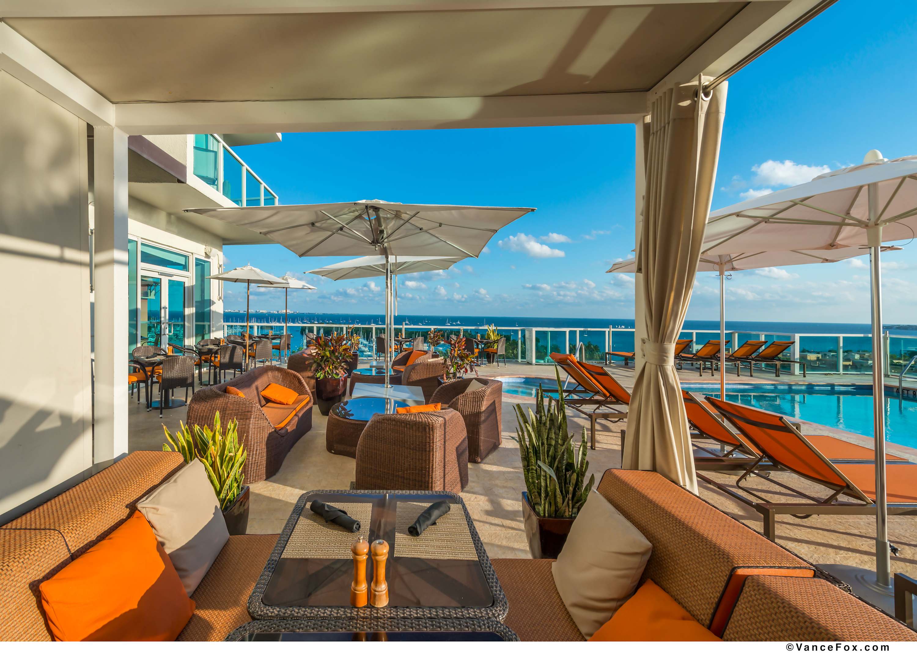 A spacious outdoor lounge area on a deck, featuring comfortable wicker sofas and armchairs with orange cushions, potted plants, and a clear view of the ocean and adjacent pool.