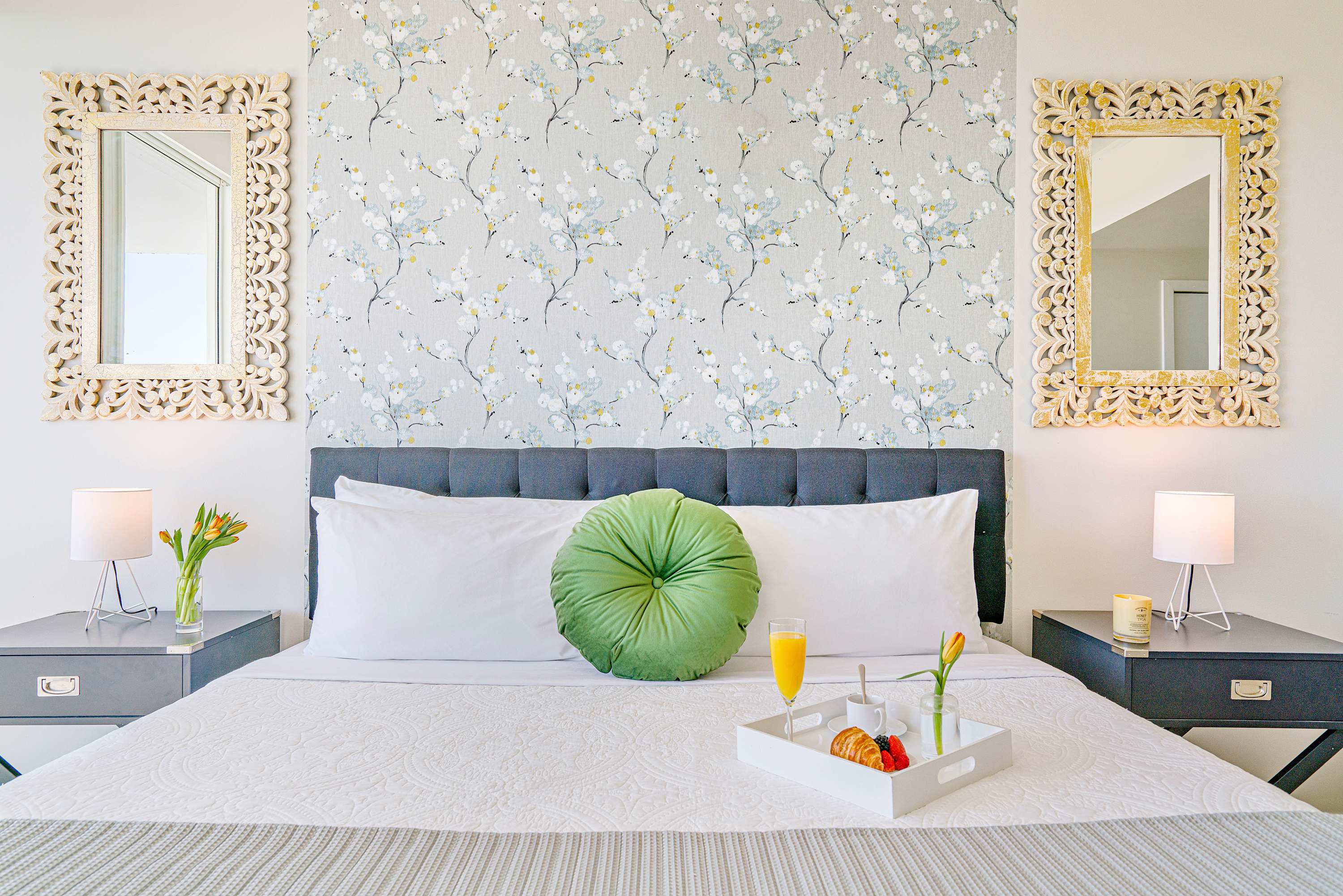 A king-sized bed with crisp white bedding and a dark upholstered headboard is centered against a light patterned accent wall, flanked by dark nightstands with lamps and ornate mirrors.