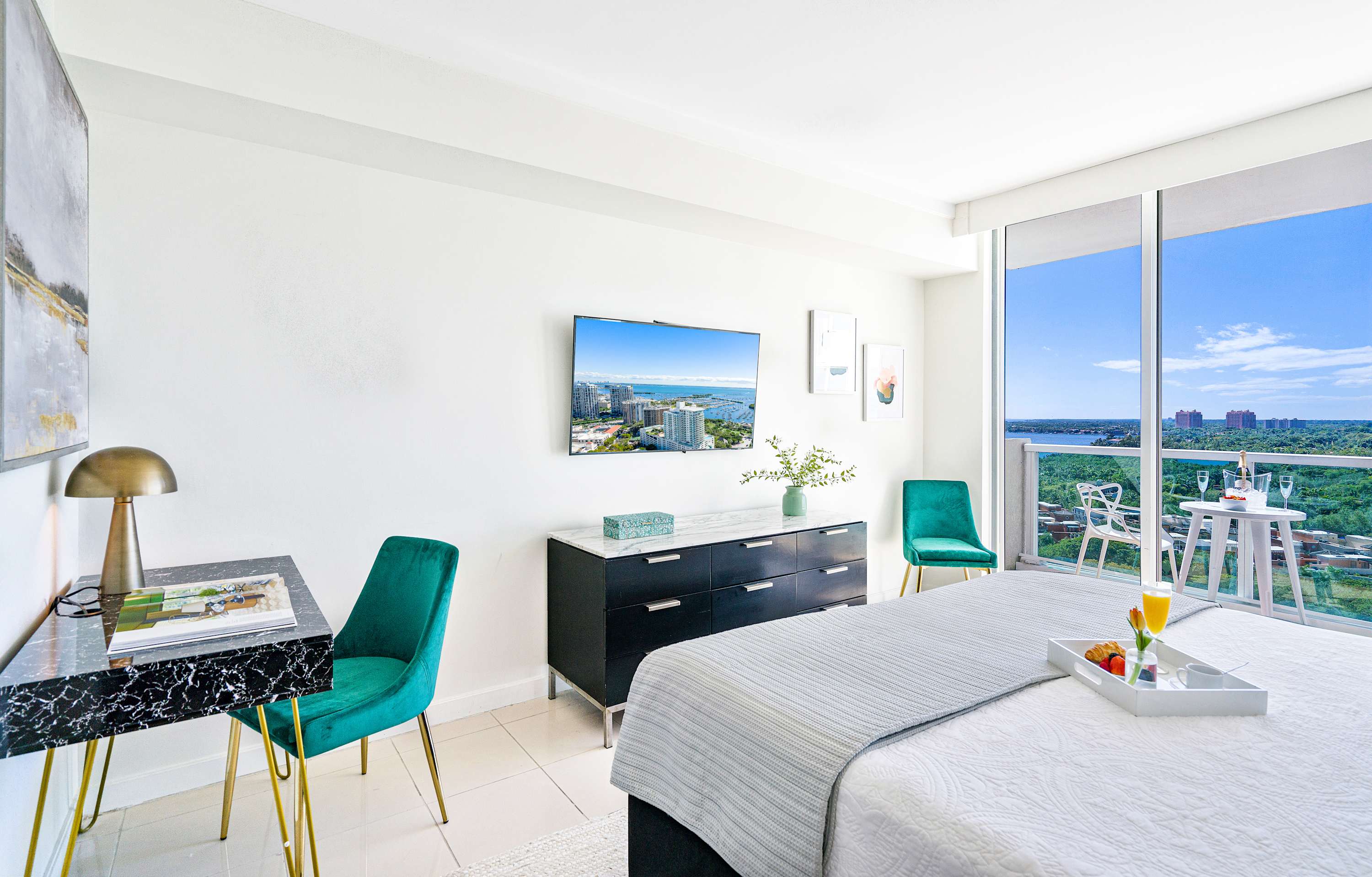 A modern bedroom features a large bed, a wall-mounted flat-screen TV displaying a city and bay view, a black dresser with a marble top, and a small desk with a teal velvet chair, all against white walls.