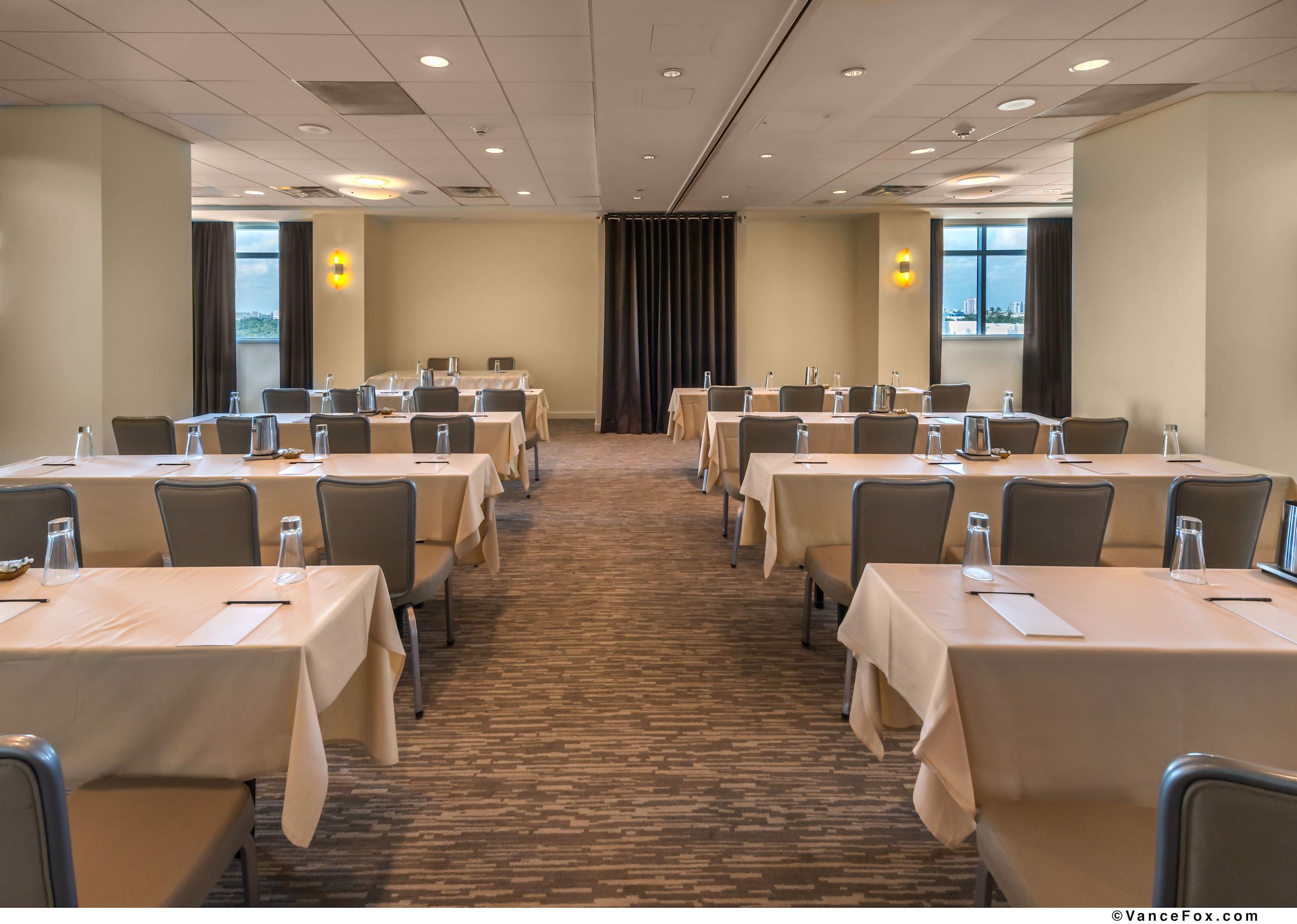 A spacious conference room is arranged with multiple rectangular tables covered in light beige tablecloths, each paired with grey upholstered chairs. The room features neutral-toned walls, patterned carpet, and windows offering glimpses of the outside, with warm accent lighting.