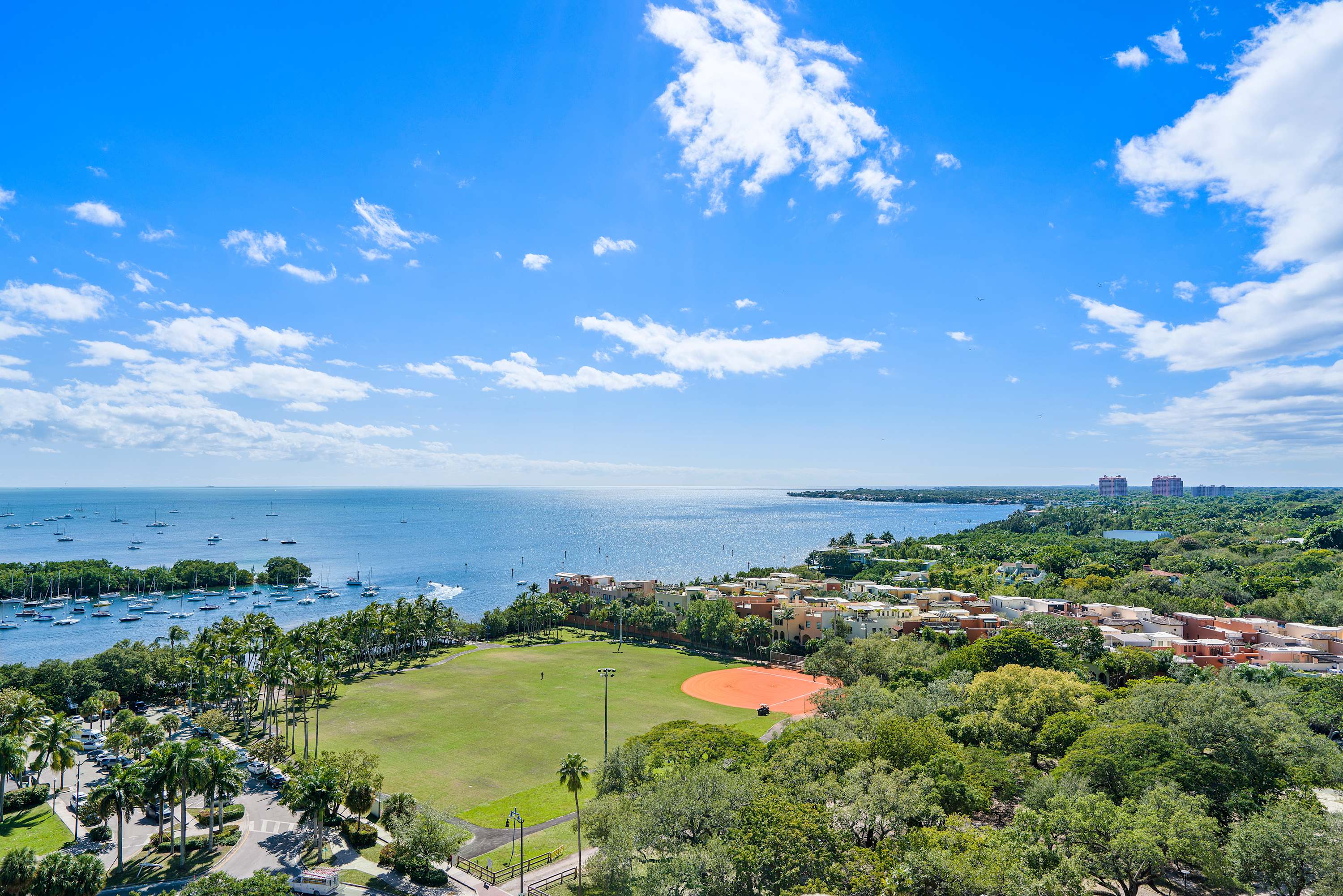 A high-angle view overlooking a green baseball field and park, a busy road, and the vast blue ocean in the distance with scattered boats under a bright sky.