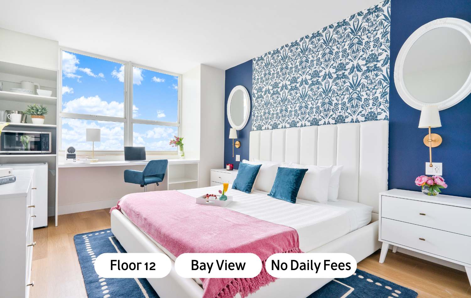Bright bedroom with a white upholstered bed, pink throw, and blue accents, featuring a large window with bay views and a compact desk area with a blue chair.
