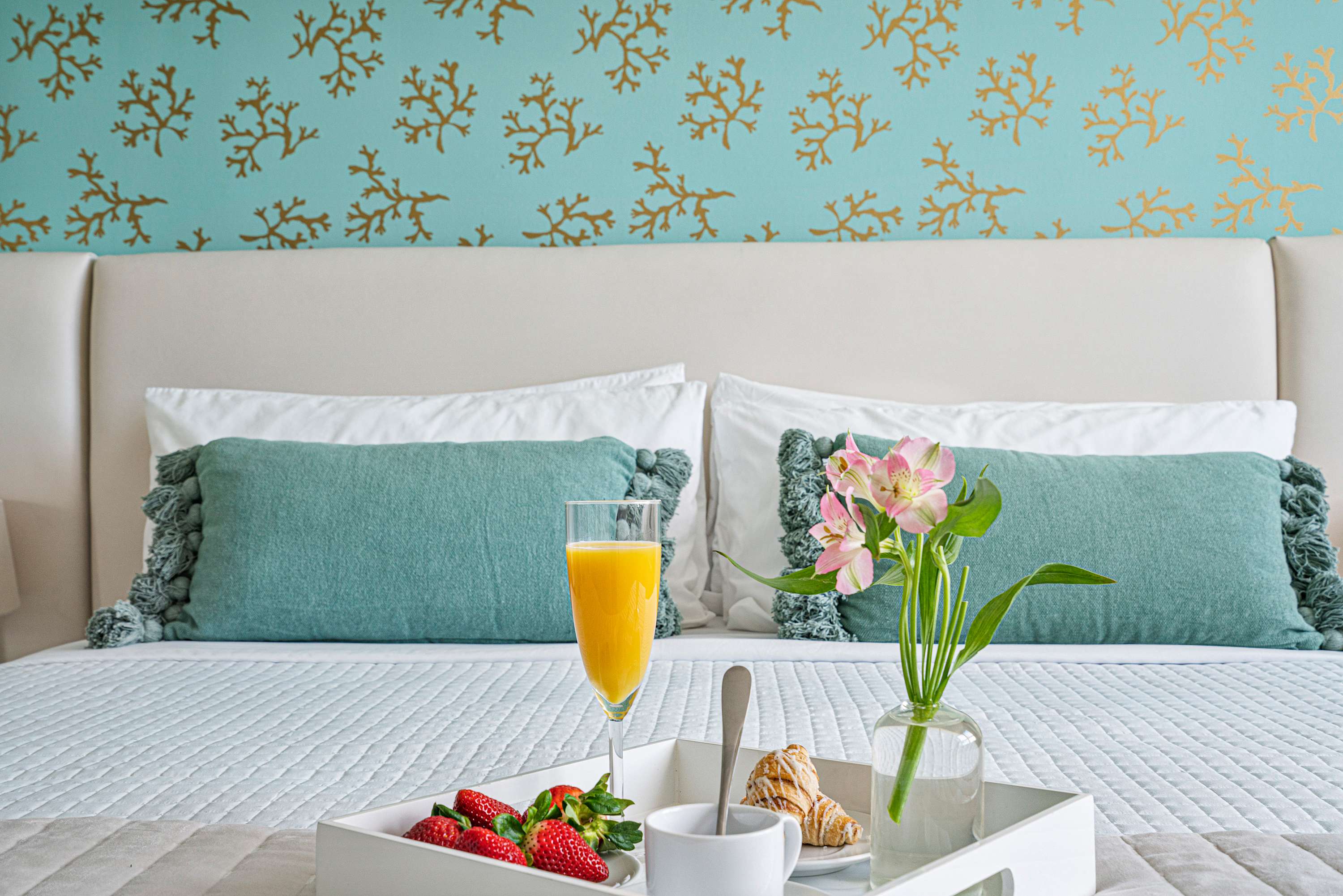 A close-up view of a white upholstered bed with crisp white linens and teal decorative pillows, featuring a breakfast tray with orange juice, strawberries, a croissant, and a vase of pink flowers. The teal and gold coral patterned accent wall is visible behind the headboard.