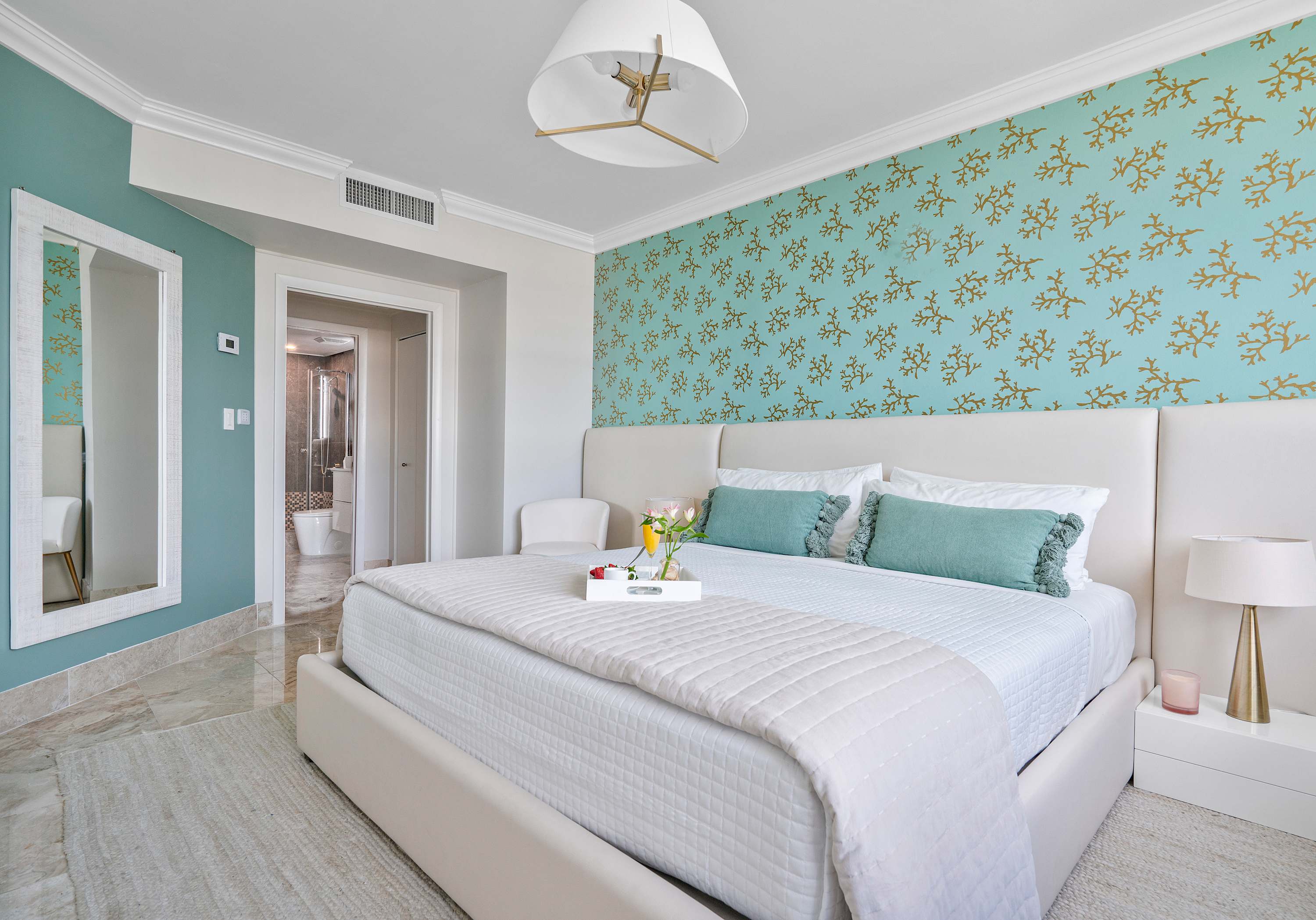 A spacious bedroom with a large white upholstered bed, white quilted bedding, and teal accent pillows, set against a striking teal and gold coral patterned accent wall. A full-length mirror with a white frame is on the left, and a glimpse of the bathroom is visible through an open doorway.