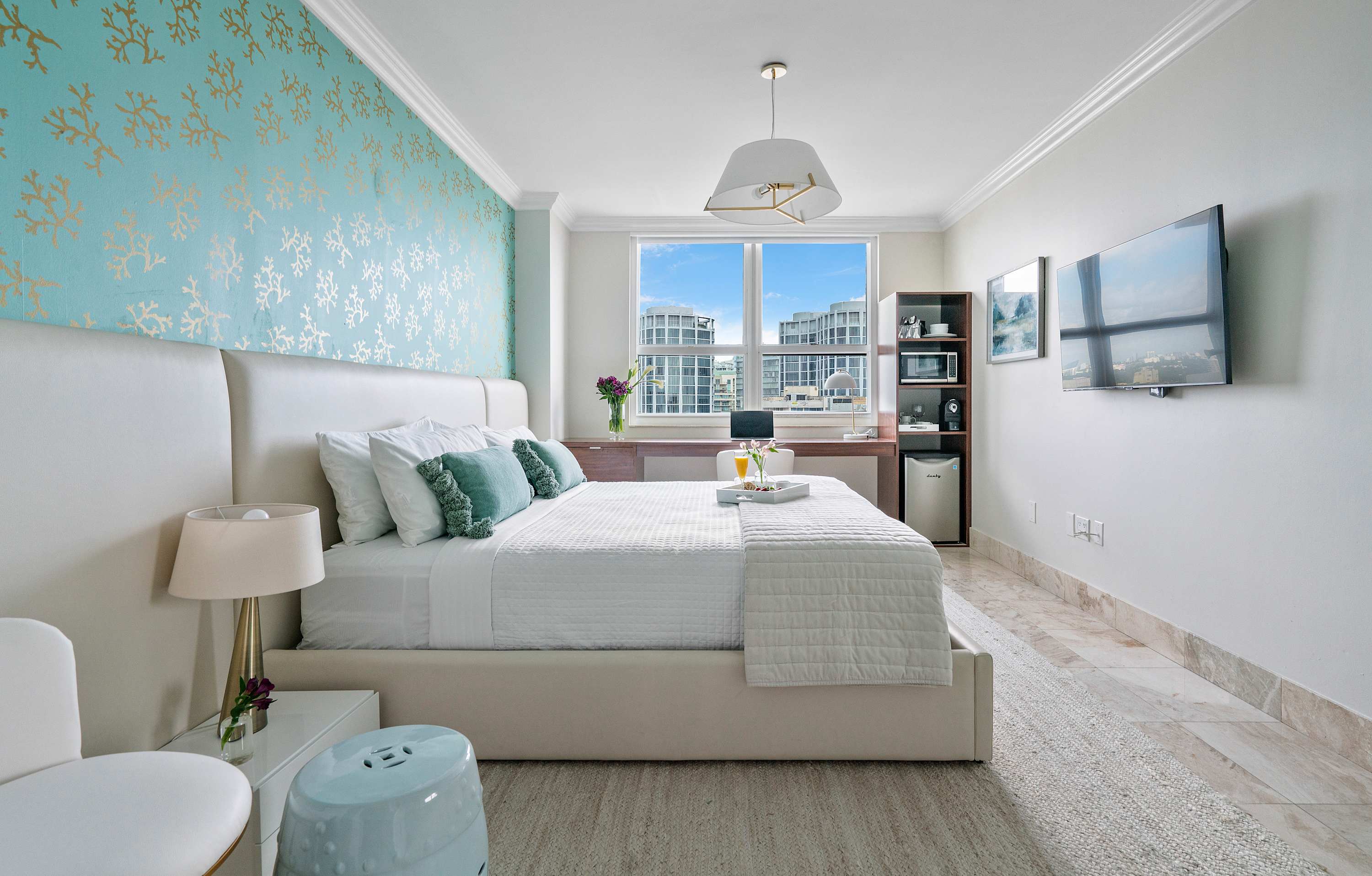 A modern bedroom with a white upholstered bed, white bedding, and teal pillows, facing a wall-mounted flat-screen TV. A long wooden desk with a white chair and a shelving unit with a microwave and mini-fridge are visible near the large window with city views.