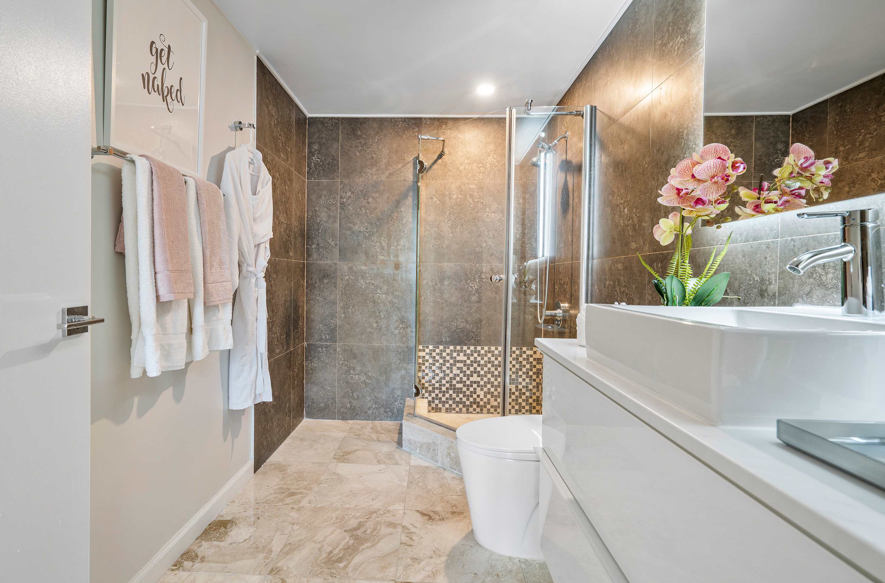 A contemporary bathroom featuring a walk-in shower with dark grey tiles and a mosaic accent, a white toilet, and a modern vanity with a vessel sink and illuminated mirror. White bathrobes and towels hang on the wall, complementing the light marble floor.