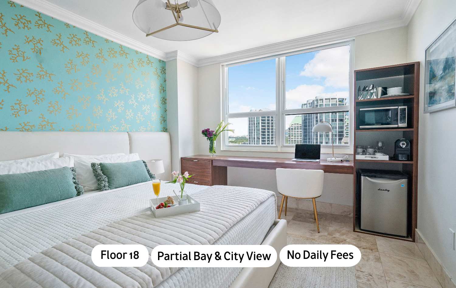 A bright bedroom featuring a white upholstered bed with teal accent pillows, a long wooden desk with a laptop and city views through a large window, and a dark wood shelving unit with a microwave and mini-fridge. The wall behind the bed has a teal and gold coral pattern.