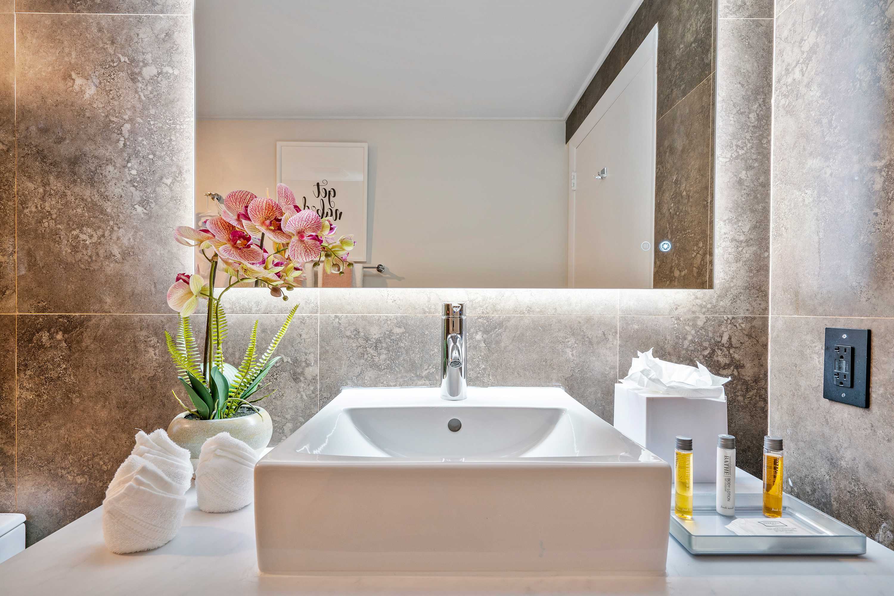 A modern bathroom vanity with a rectangular vessel sink, a large illuminated mirror, and a white countertop. Decorative elements include rolled towels, a potted orchid, and small amenity bottles, all set against textured grey tile walls.