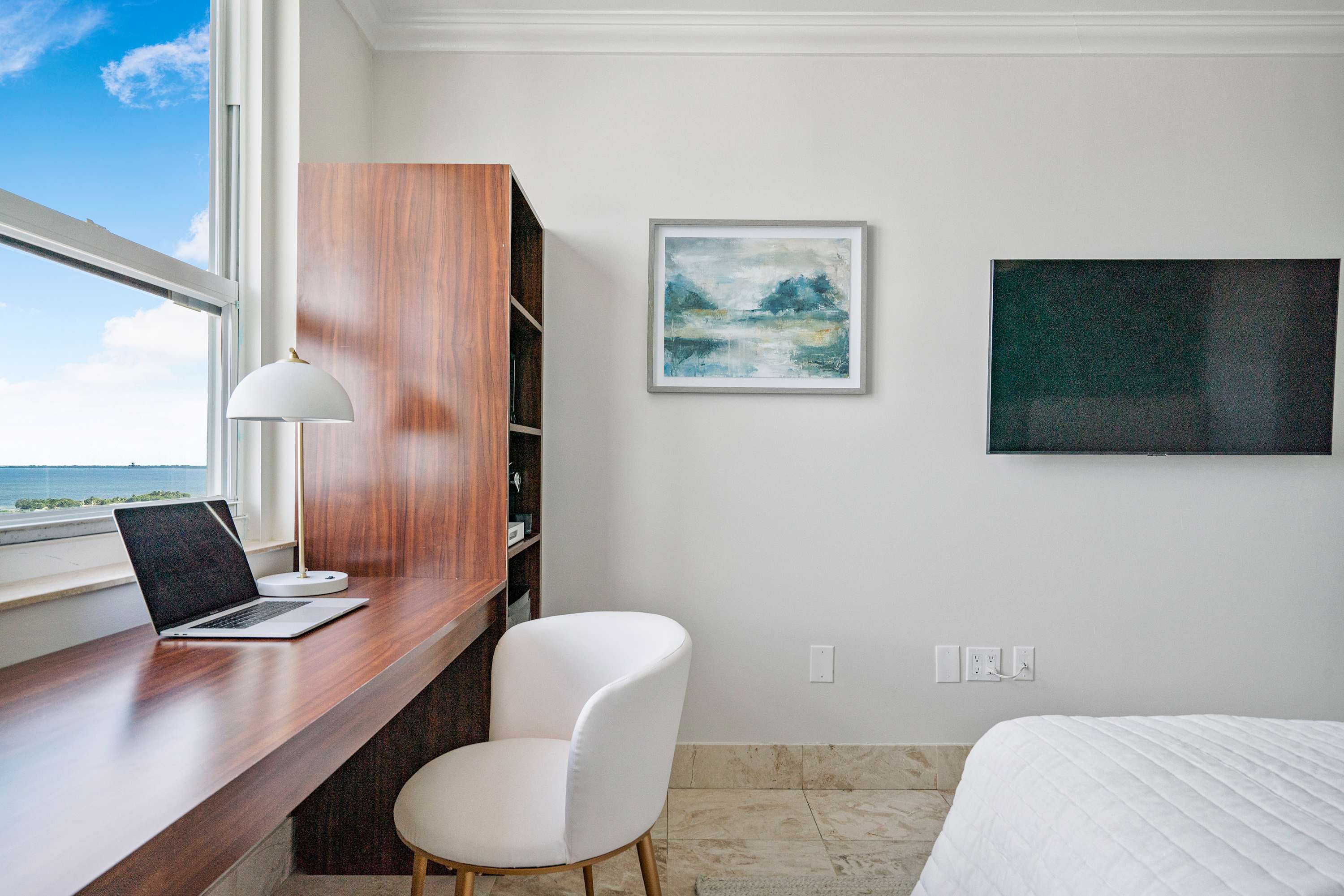 A dedicated workspace within the bedroom, featuring a sleek wooden desk with a laptop and a white modern chair, positioned by a large window offering partial bay and city views. A dark wood shelving unit is integrated next to the desk, and a framed abstract painting hangs on the wall.