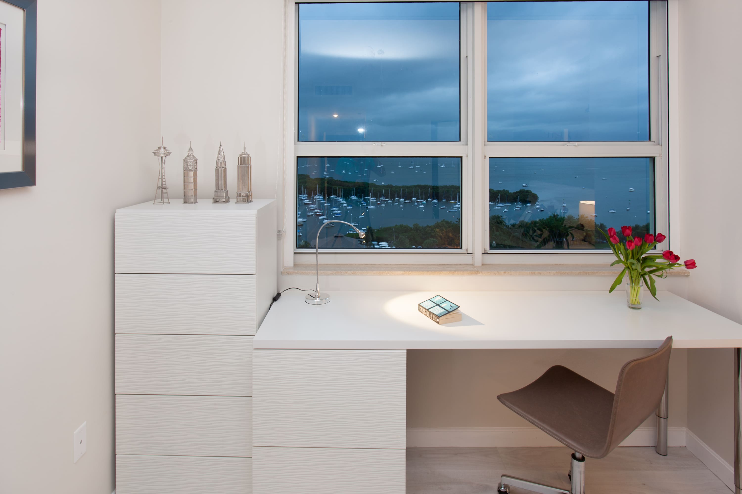 Modern white desk with a brown office chair positioned by a large window, offering a partial view of the bay with boats. A white dresser with decorative city landmarks sits next to the desk.