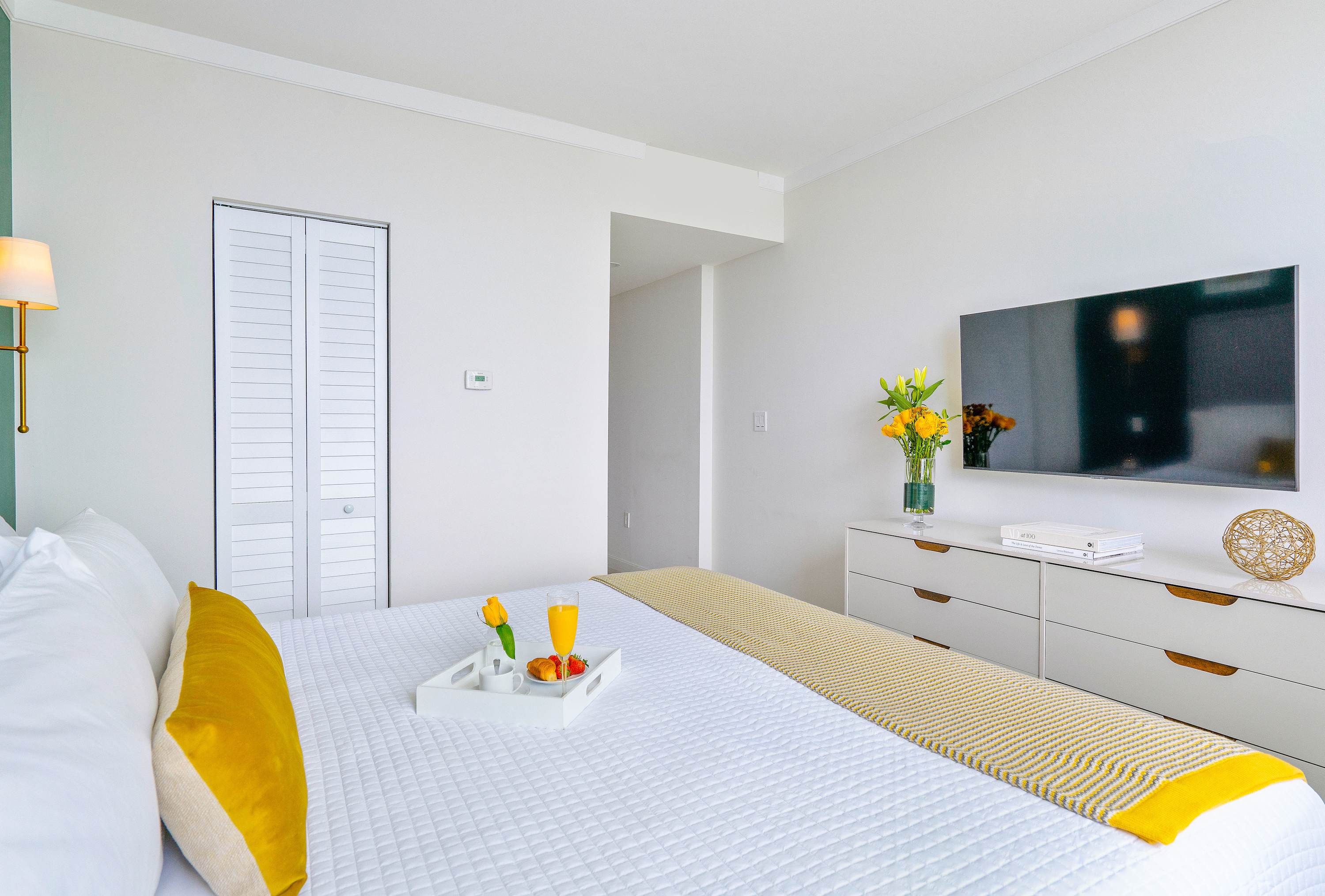 A modern bedroom with a queen bed, white textured bedding, and a yellow throw, facing a wall-mounted flat-screen TV above a white dresser. A white bi-fold closet door is visible, and a tray with breakfast items rests on the bed.