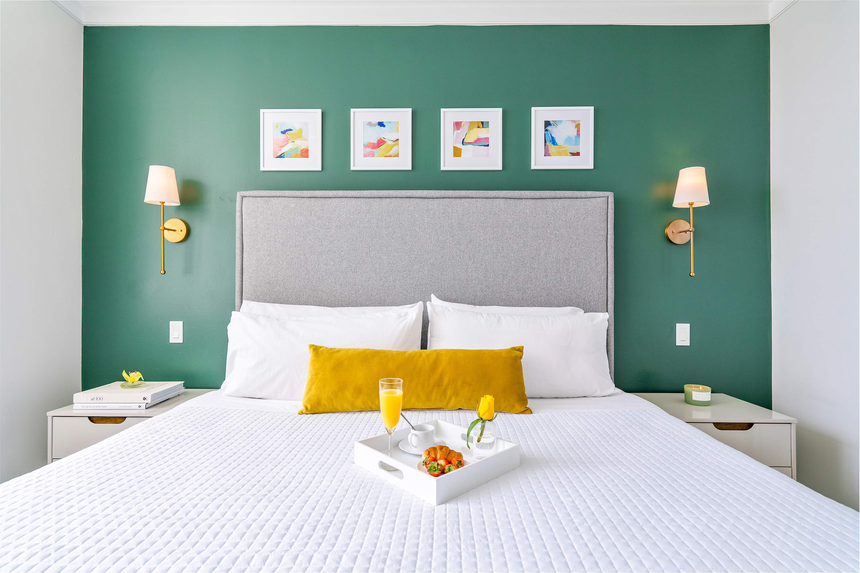 A comfortable queen bed with a gray upholstered headboard, crisp white bedding, and a yellow throw pillow, set against a vibrant green accent wall adorned with four framed abstract artworks. Gold wall sconces flank the bed, providing warm lighting.