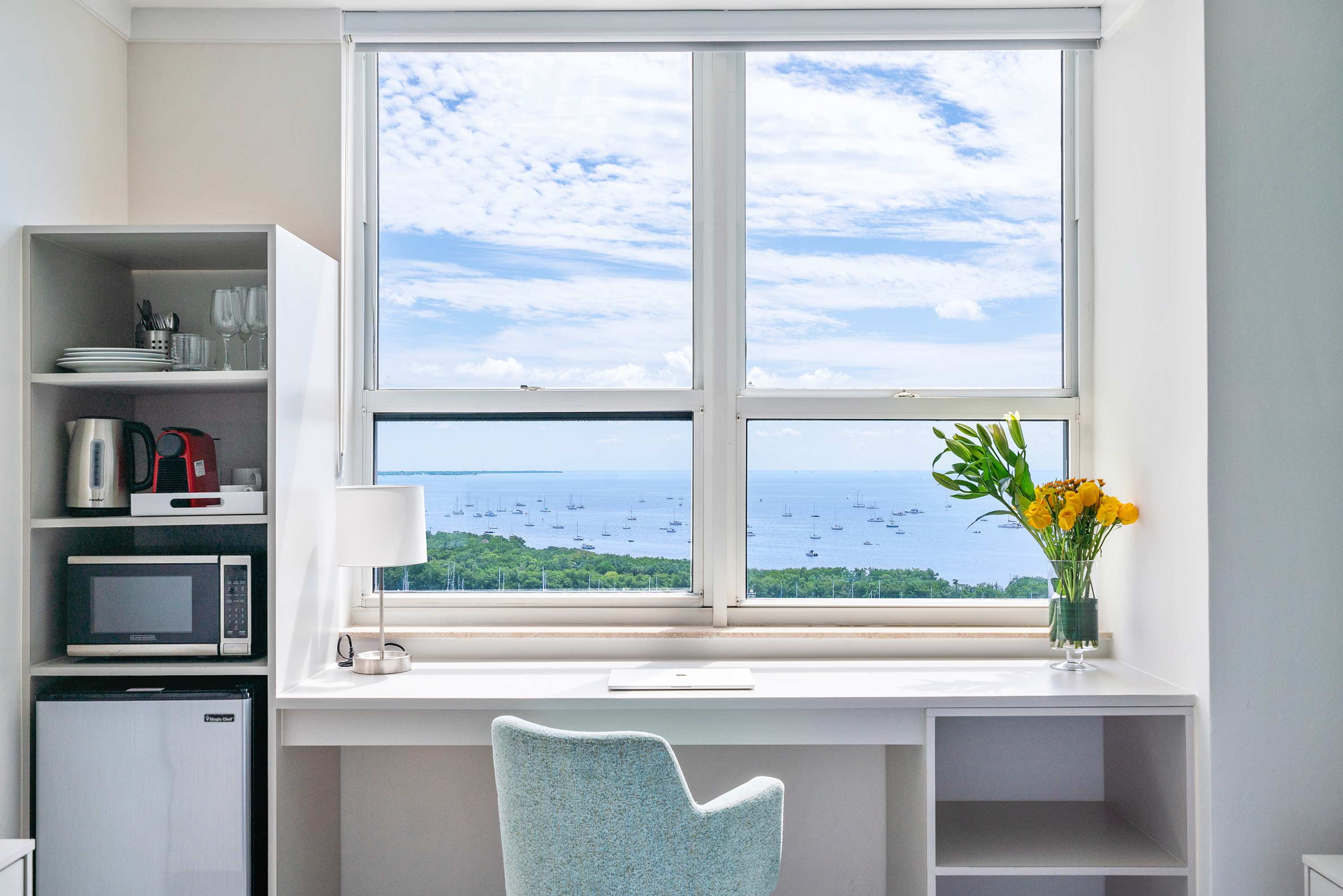 A well-lit studio space featuring a white built-in desk with a light blue chair positioned by a large window offering expansive bay views, alongside a compact kitchenette with open shelving, a microwave, and a mini-fridge.