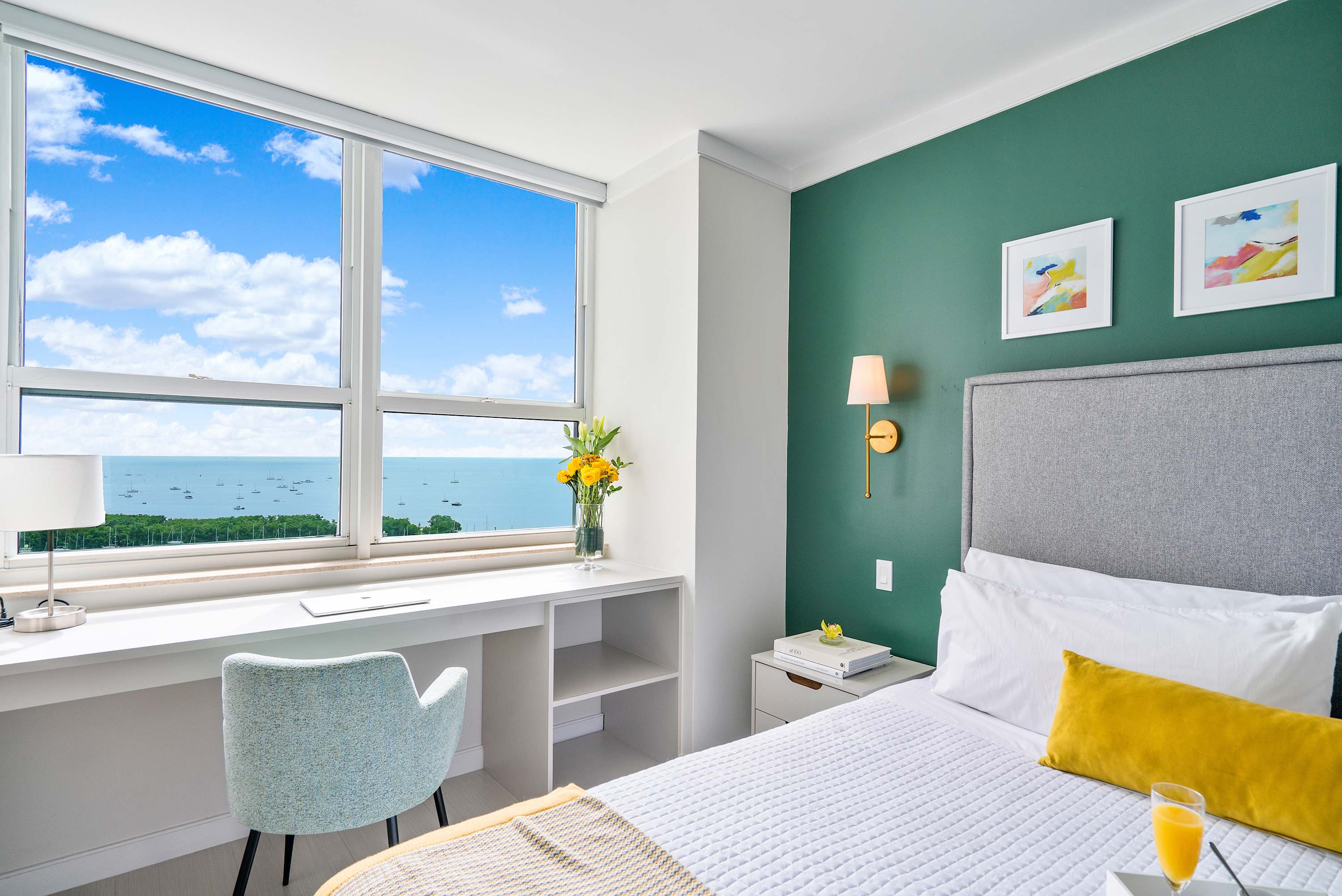 A bright bedroom featuring a queen bed with a gray upholstered headboard, a built-in white desk and chair by a large window offering bay views, and a vibrant green accent wall. The room is decorated with modern art and gold wall sconces.