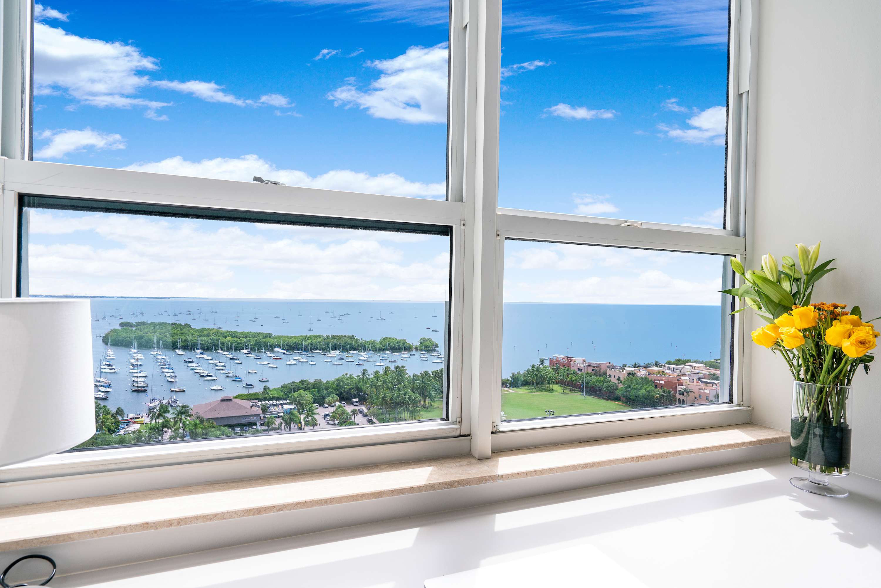 A stunning bay view through a large window, showcasing a marina filled with numerous boats, lush green islands, and distant coastal buildings under a bright blue sky with fluffy white clouds. A vase of yellow flowers sits on the windowsill.