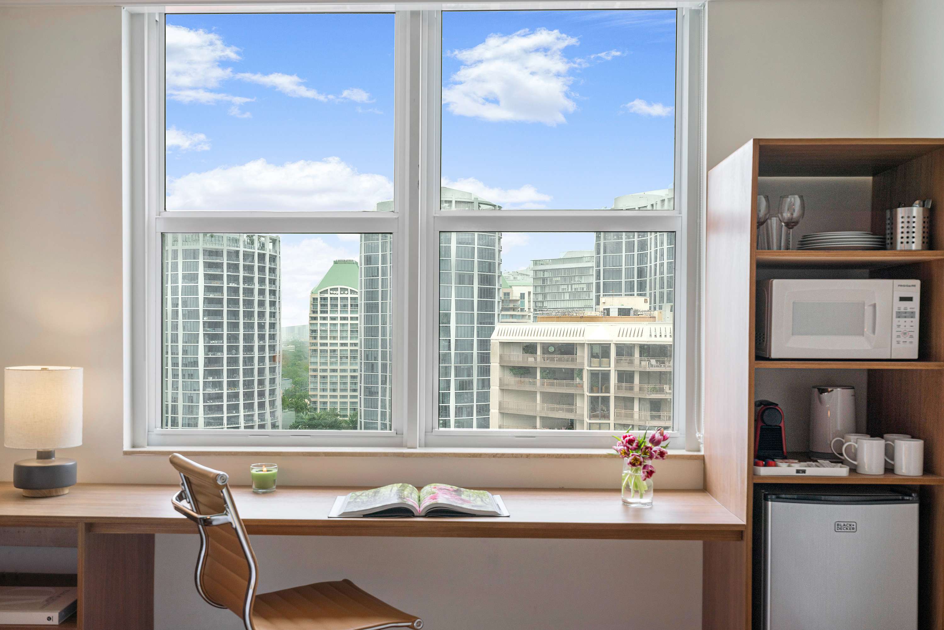A dedicated workspace with a light wood desk and chair, positioned beneath a large window offering a partial city view. A mini-fridge, microwave, and coffee maker are neatly arranged on a shelving unit beside the desk.