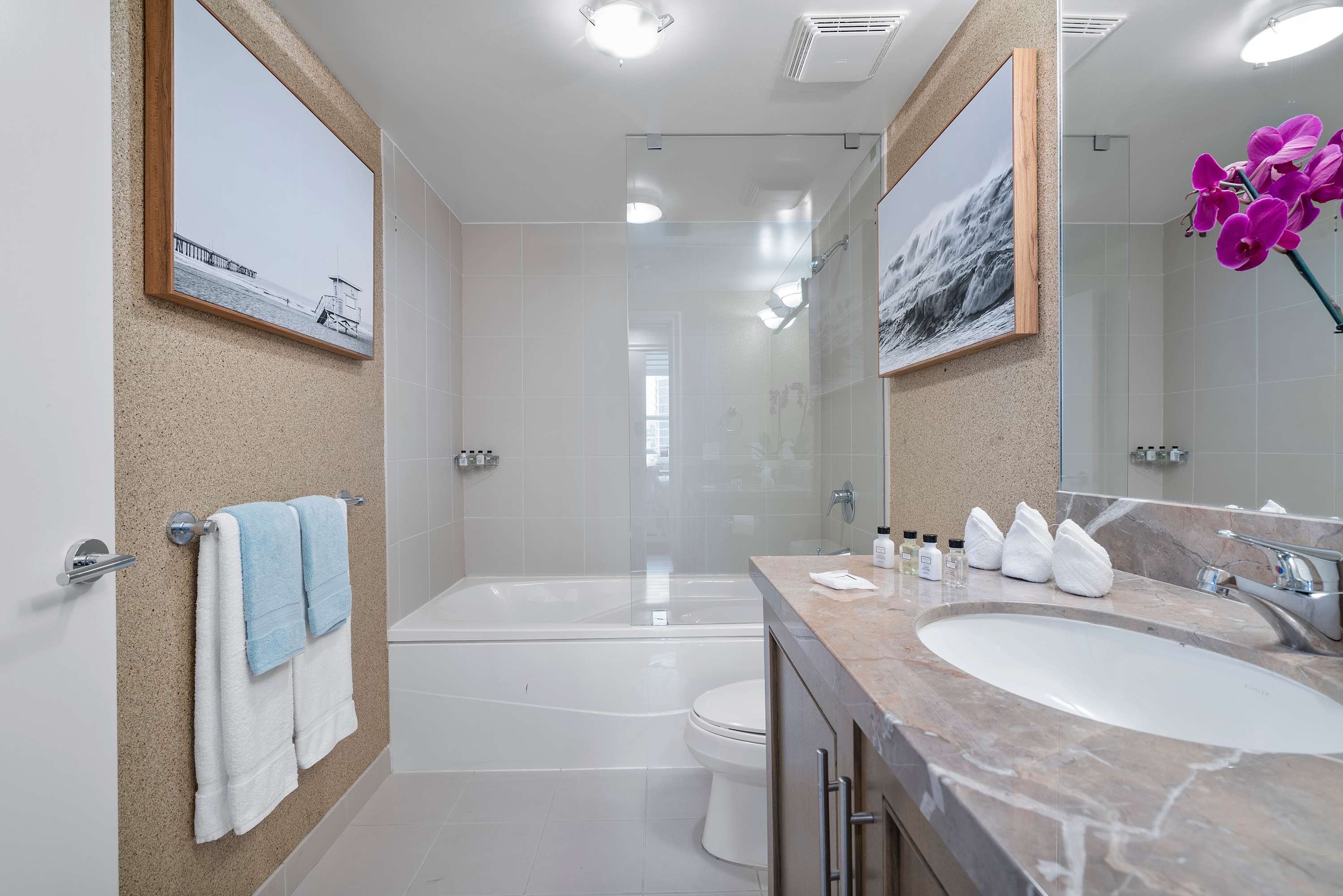 A clean and bright bathroom with a single vanity, marble countertop, and a large mirror. The shower/tub combination is visible, featuring light-colored tiles and a glass partition.