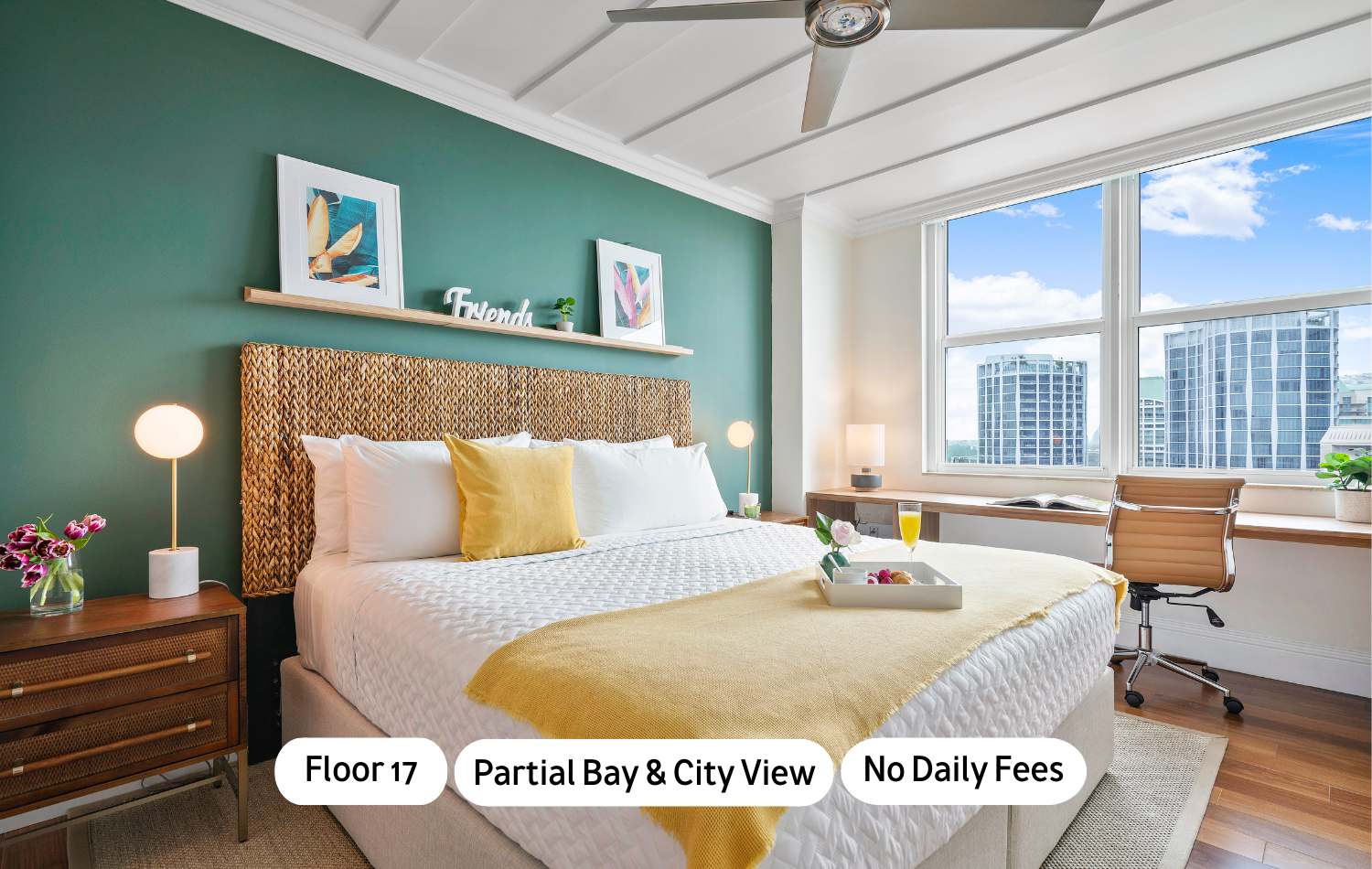 A bright bedroom featuring a queen-sized bed with a woven headboard, yellow accents, and a teal accent wall. A desk with a chair is positioned by a large window offering city views.