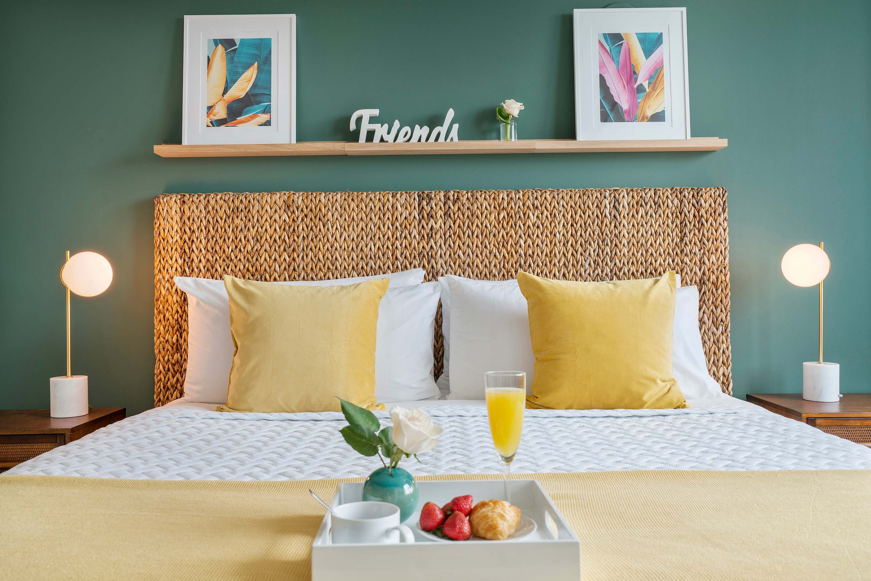 Close-up of a queen-sized bed with a textured woven headboard, white bedding, and yellow throw pillows. A breakfast tray with juice and fruit rests on the bed.