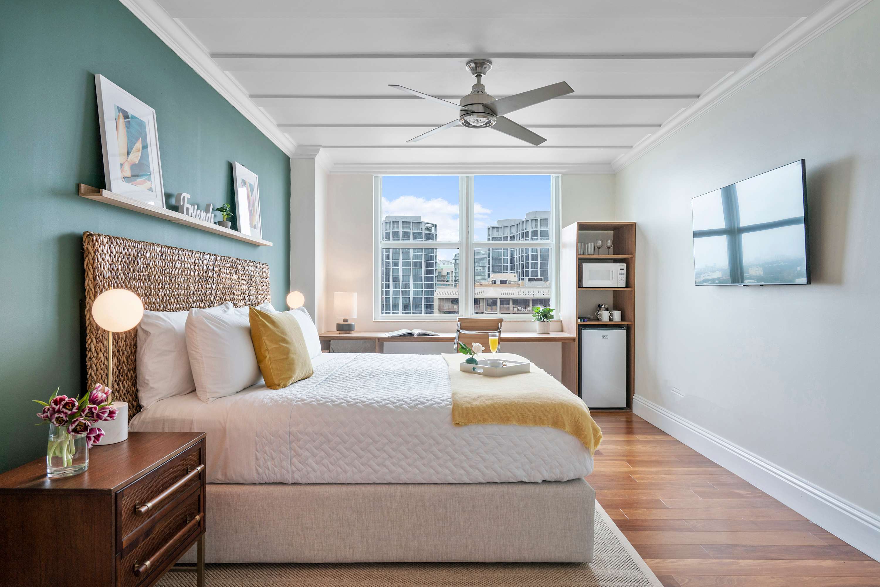 A spacious bedroom with a queen bed, a teal accent wall, and a desk by a large window offering city views. A wall-mounted TV and a shelving unit with a microwave and mini-fridge are also visible.