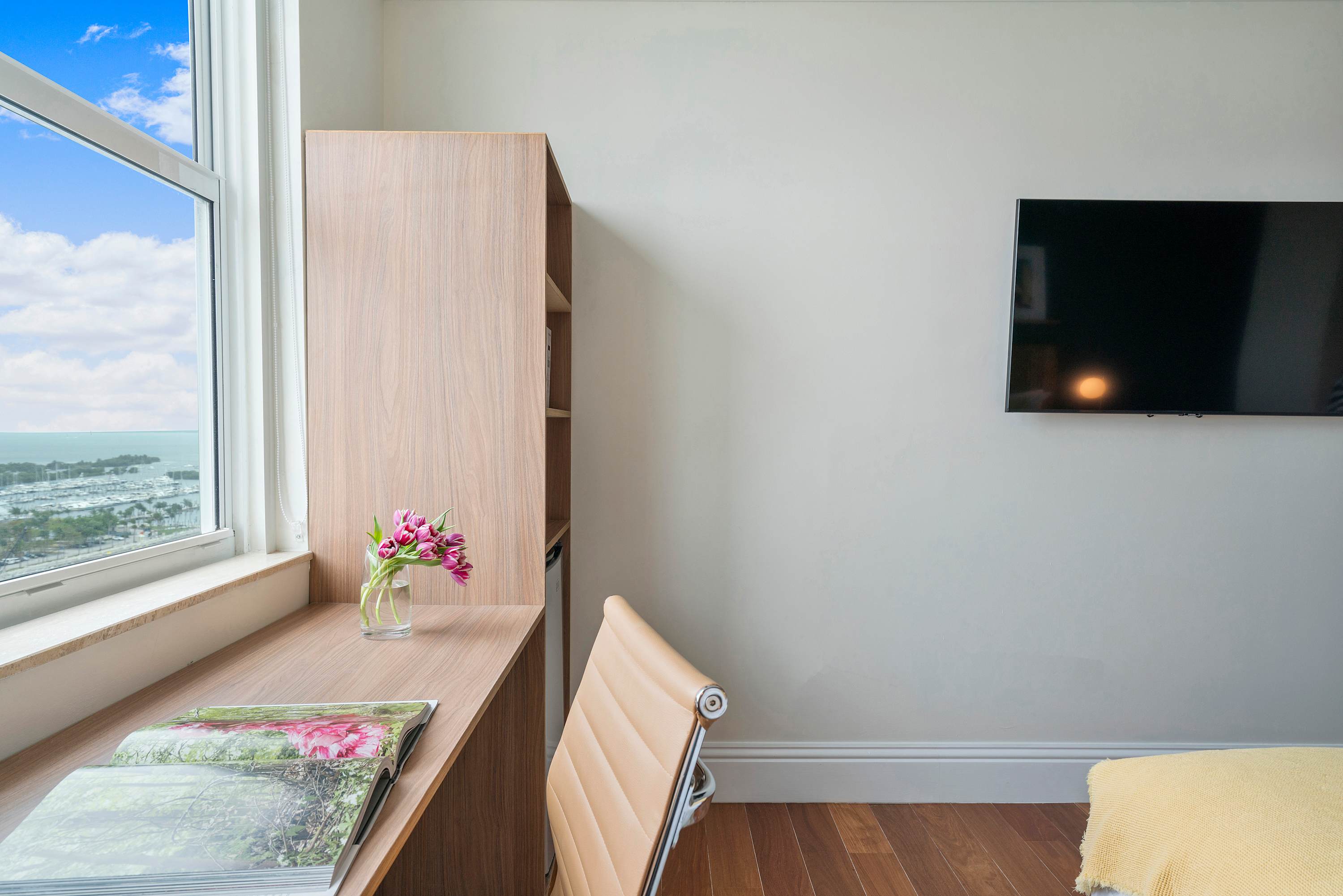A bright corner of the bedroom featuring a light wood desk by a large window with a partial bay view. A wall-mounted flat-screen TV is visible on the adjacent white wall.