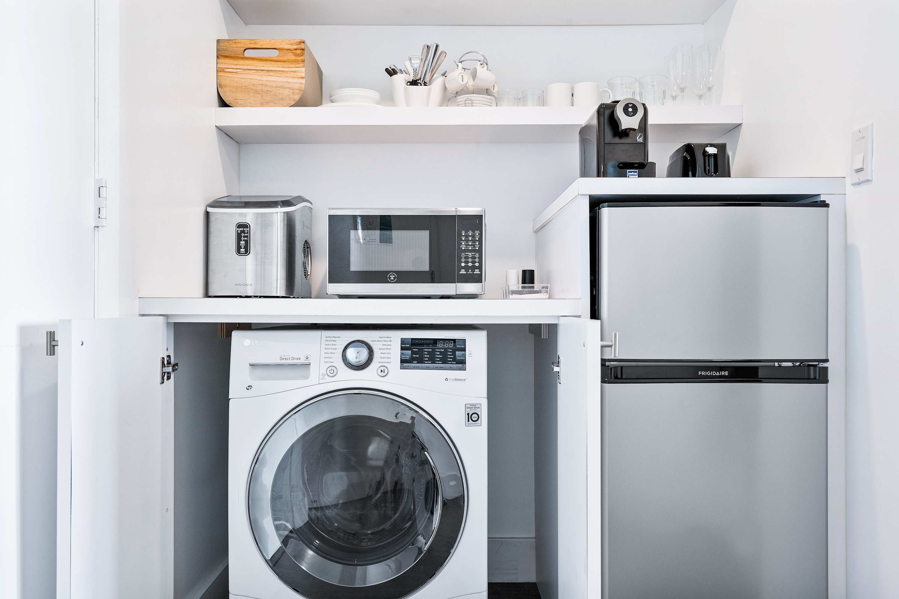 A compact laundry and kitchenette area features a stacked washer/dryer unit, a mini-fridge, microwave, ice maker, and coffee machine, all neatly integrated into white cabinetry with open shelving.